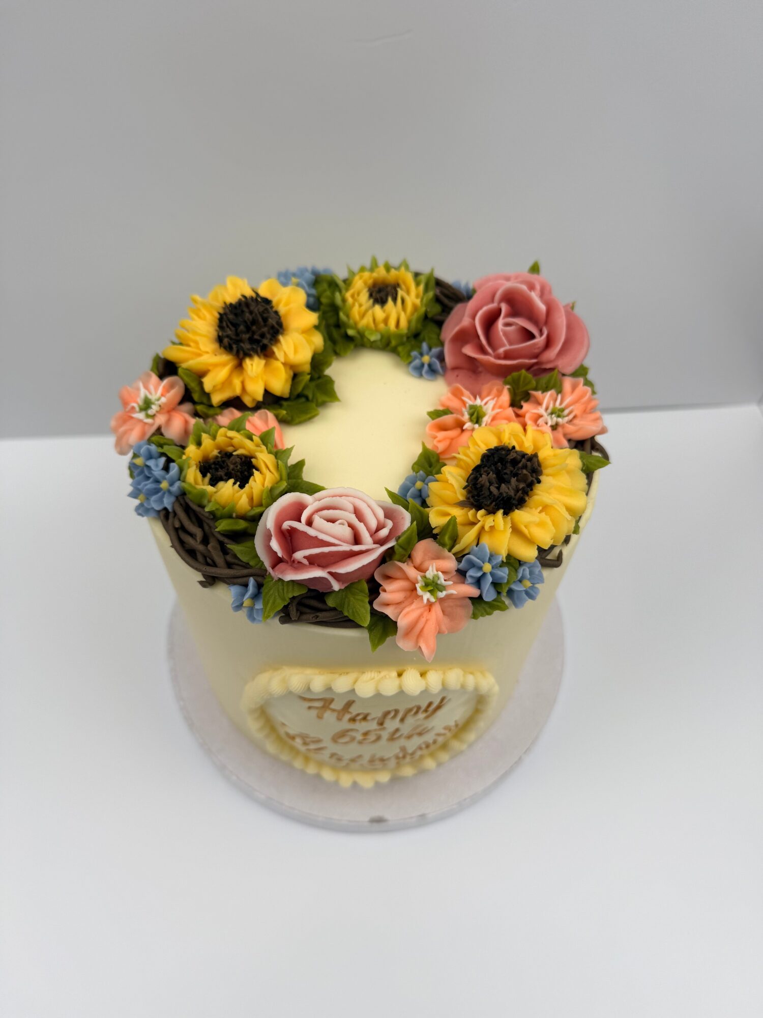 Decorative cake with sunflower, pink roses, and small pink and blue flowers, topped with a message, on a white surface.