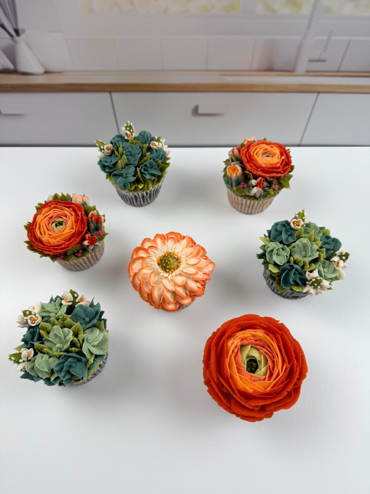 Seven cupcakes decorated with colorful flowers and a central pumpkin-shaped cupcake on a white surface.