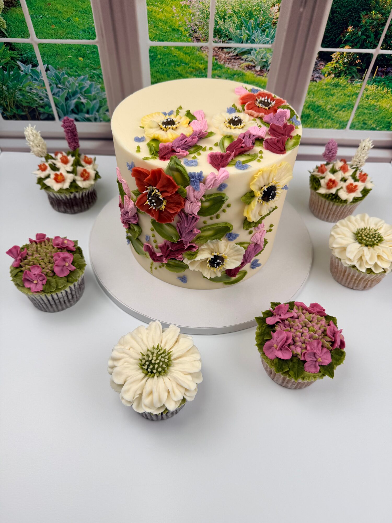 Round cake decorated with colorful flowers, surrounded by six cupcakes with floral designs, on a glass table near windows.