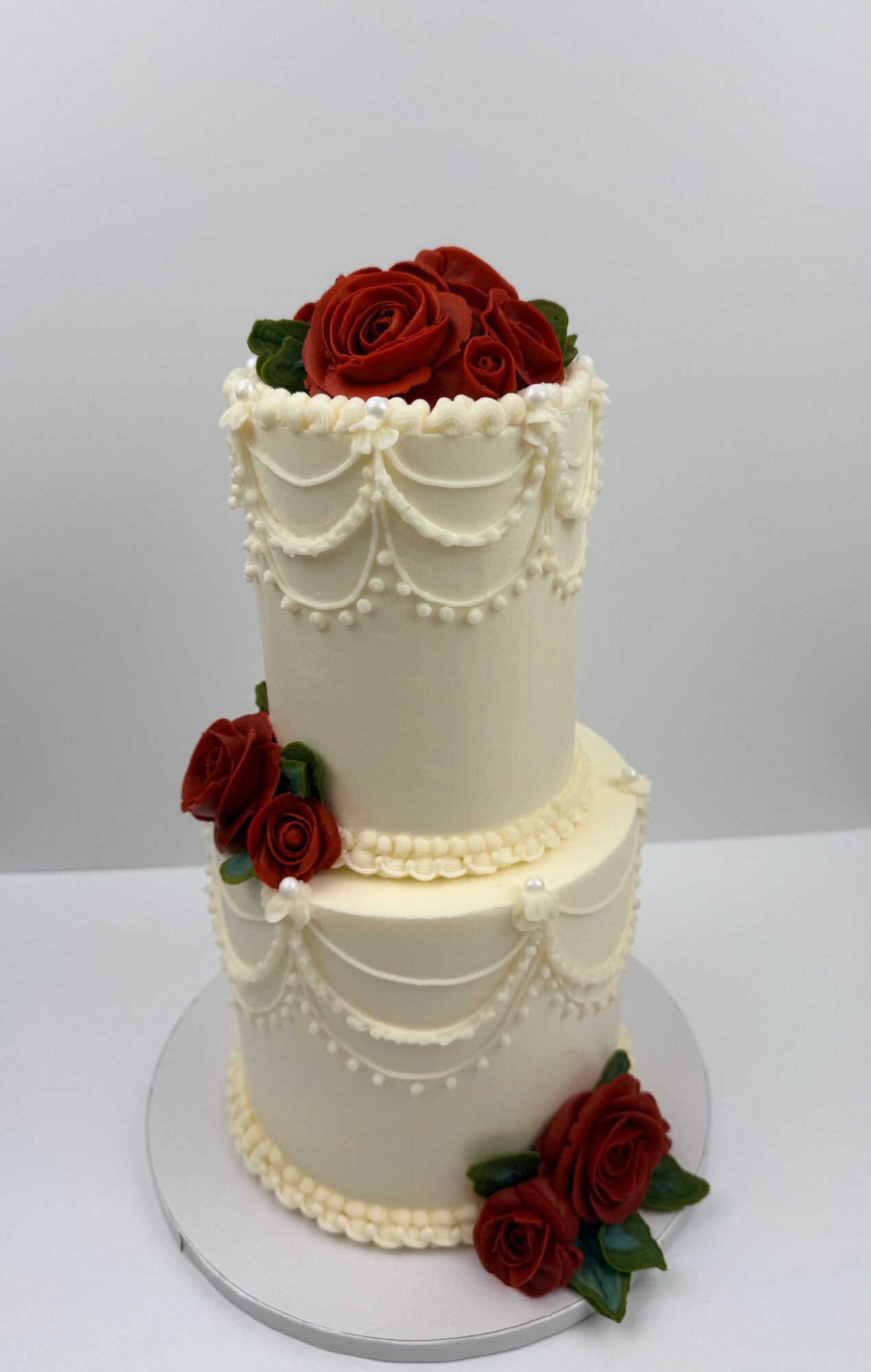 Two-tiered white cake decorated with red roses and pearl-like icing patterns, with a plain background.