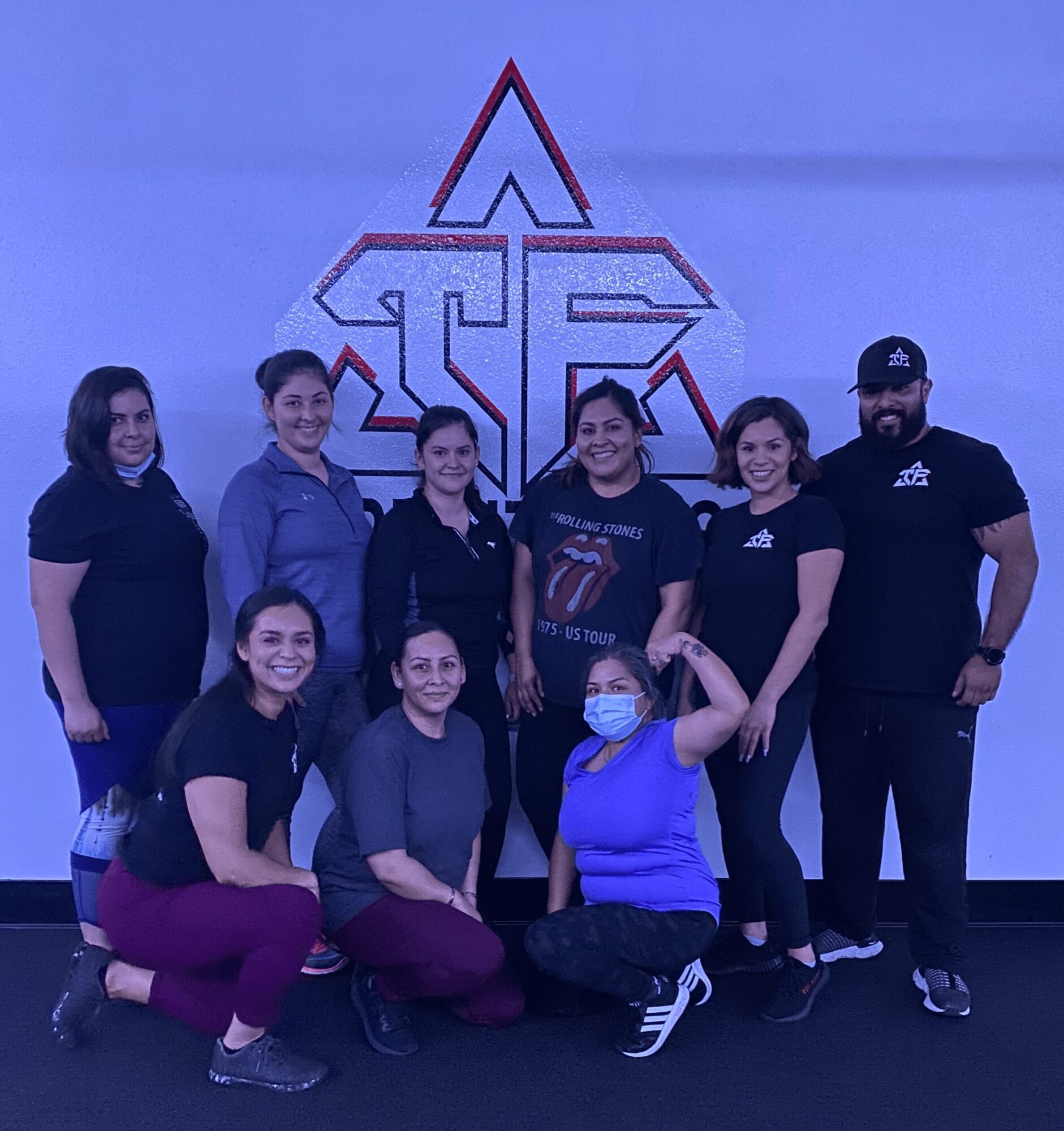 Group of nine people, some wearing masks, standing and sitting in front of a geometric logo on a wall.
