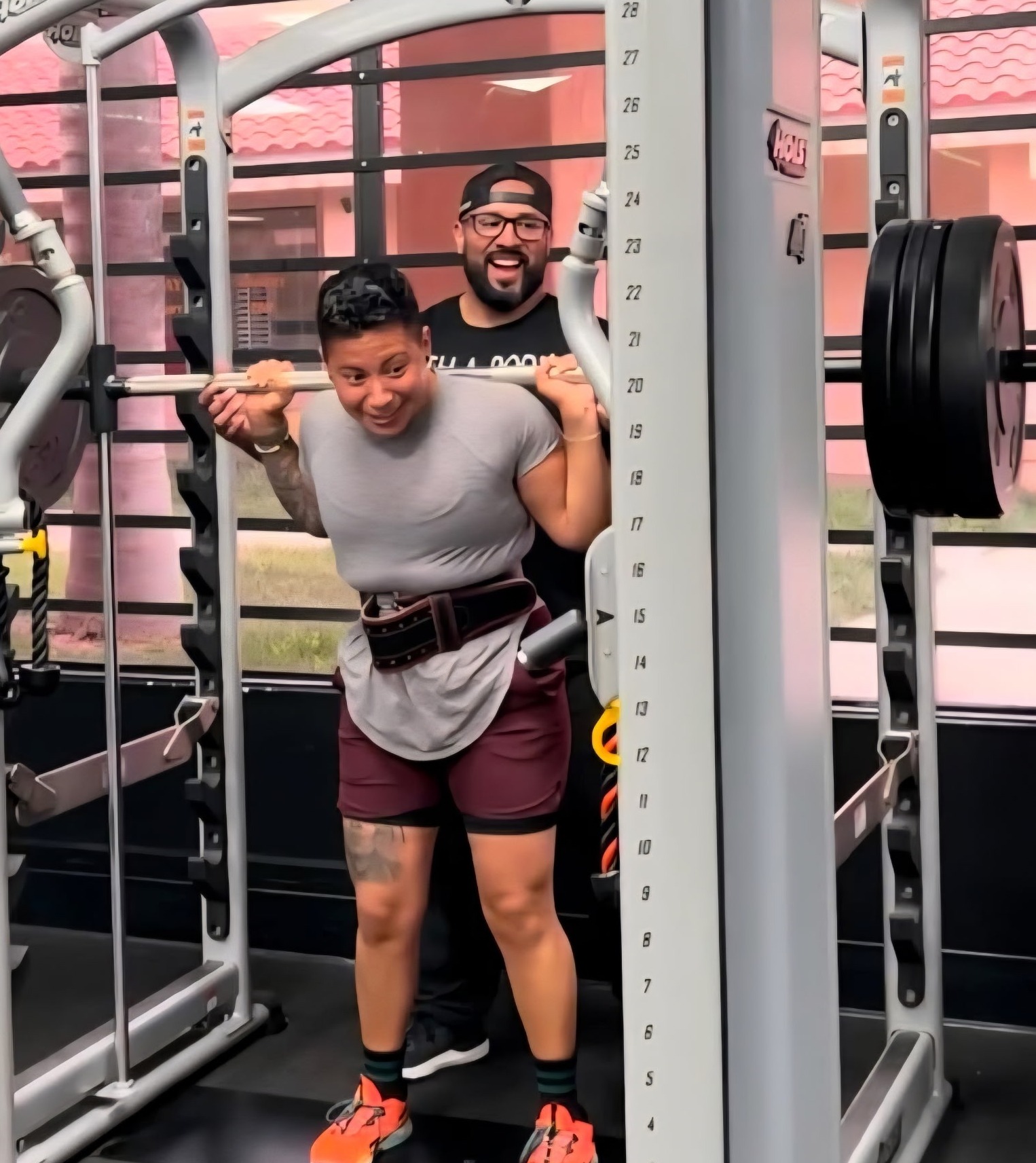 Two people in a gym, one lifting weights and the other smiling behind, with gym equipment and windows in the background.