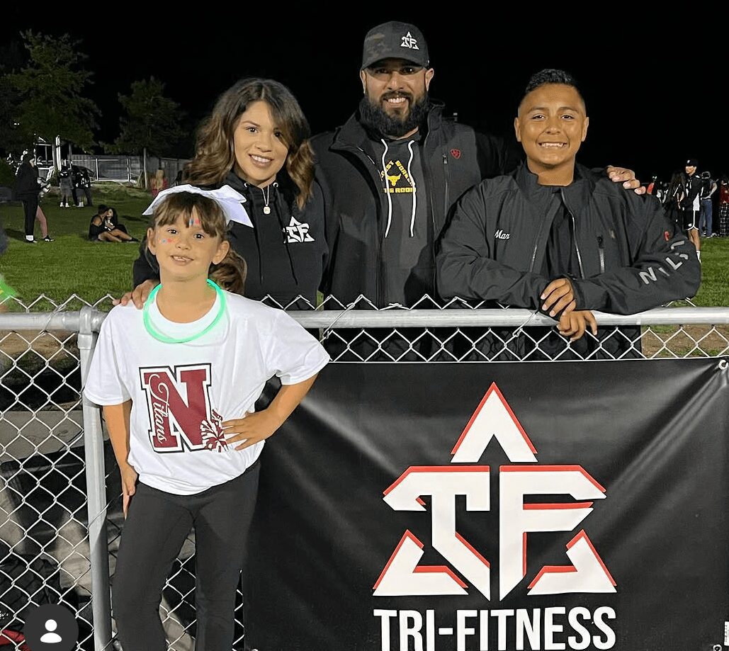 Four people, two children and two adults, pose behind a fence at night, with a Tri-Fitness banner in front.