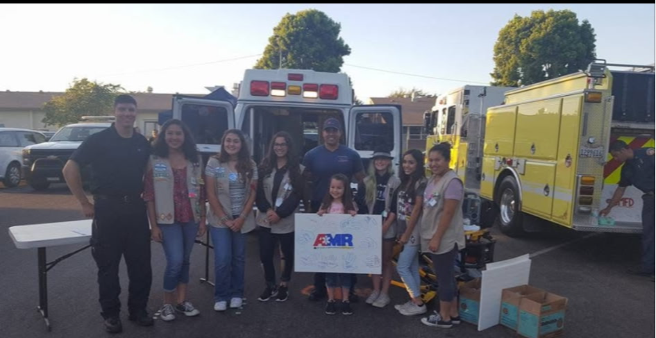 Group of people standing in front of emergency vehicles, holding a sign, outdoors on a sunny day.