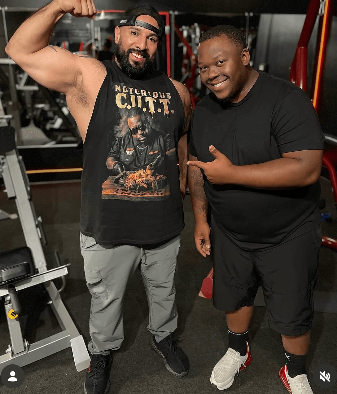 Two men smiling and posing in a gym, one flexing arm muscle, the other pointing at him, gym equipment in background.