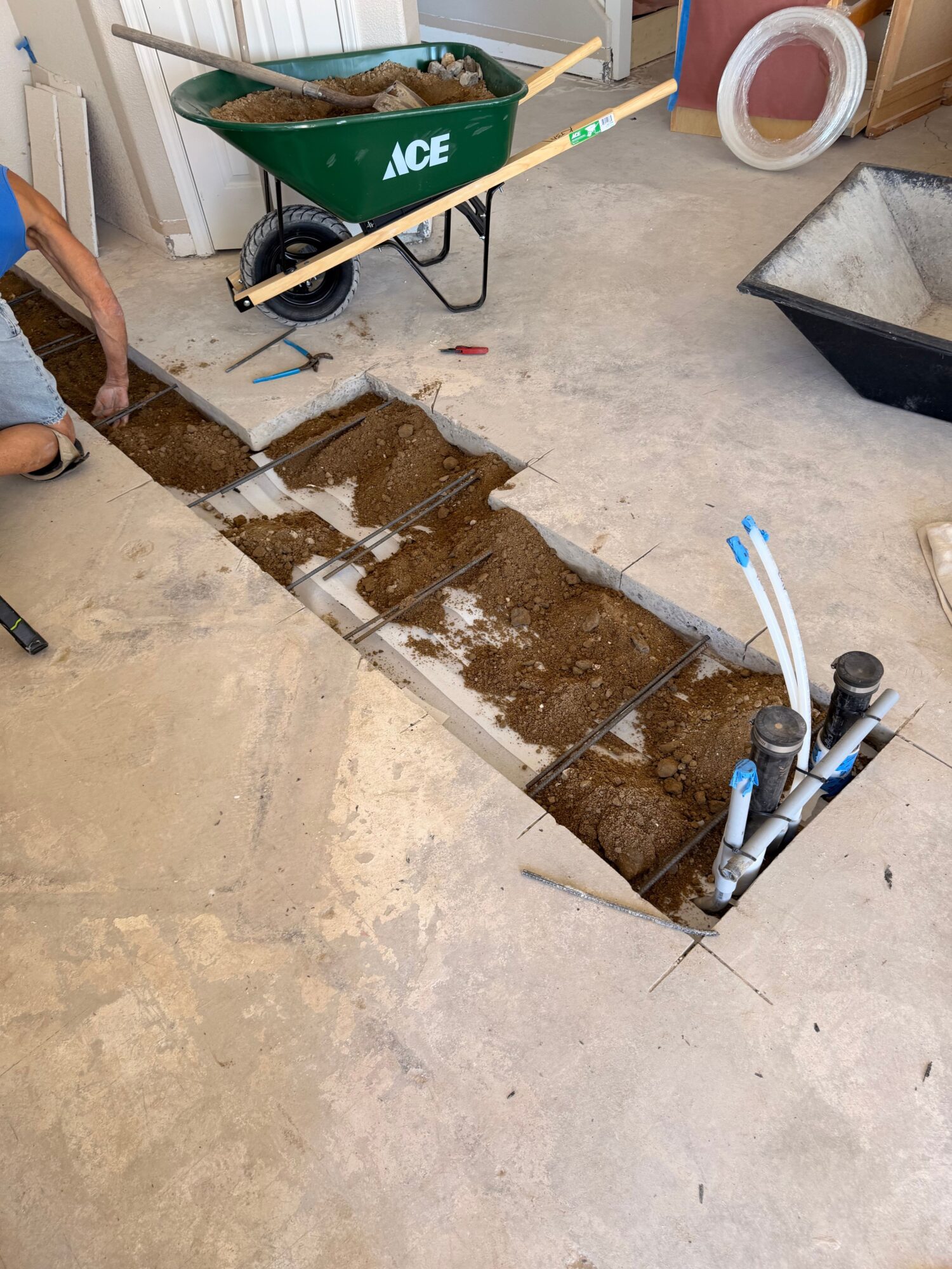 Open floor pipe access with exposed plumbing and dirt, surrounded by construction tools and materials.
