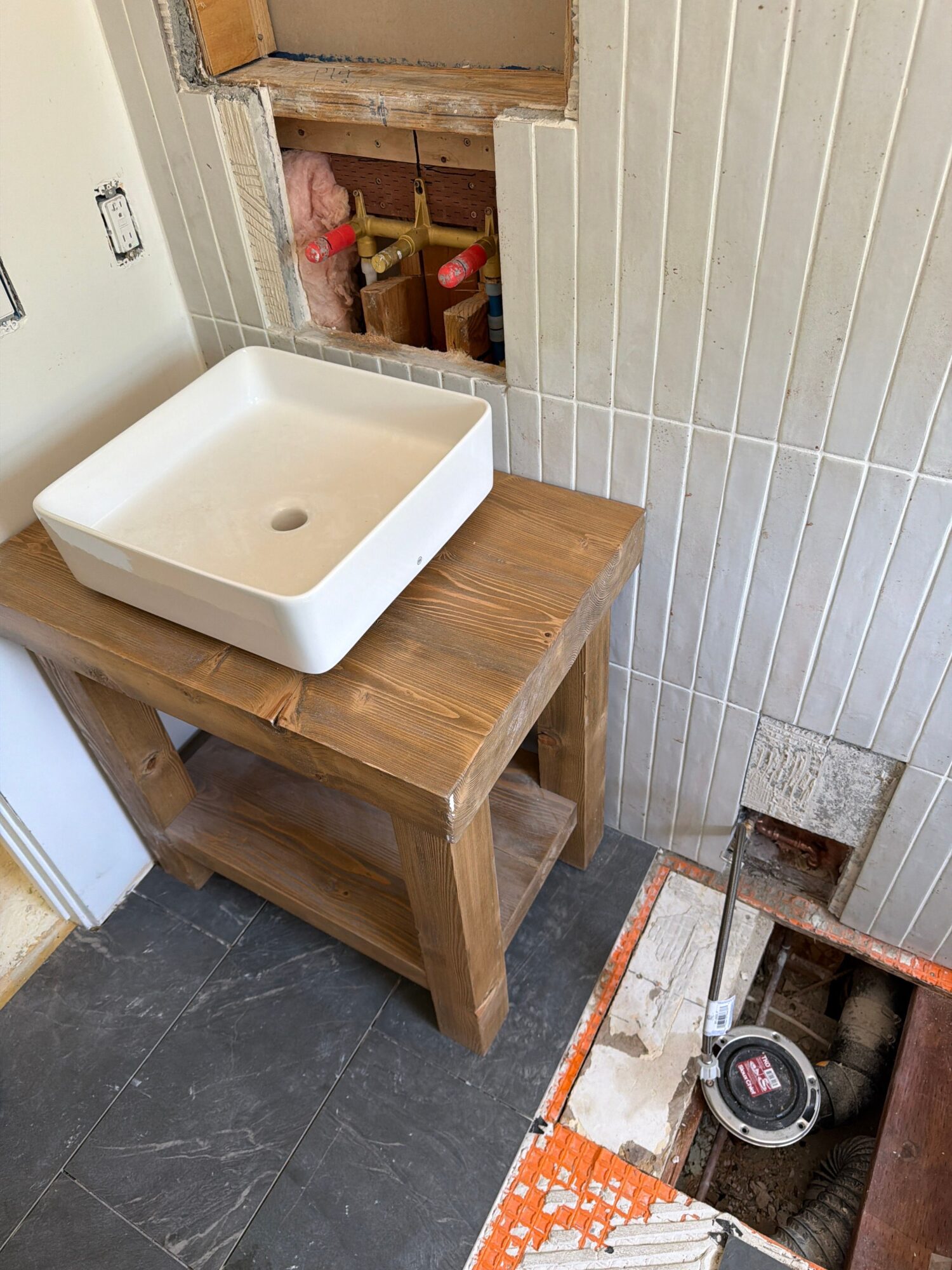 Wooden sink vanity with a white basin, against a tiled wall and dark floor, under construction.