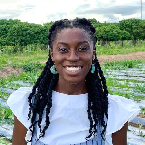 Young woman with braided hair smiling outdoors in a field with water and green trees behind her.