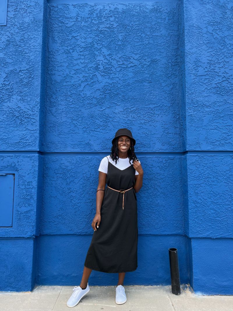 Woman standing against a textured blue wall, smiling, wearing a black dress, white t-shirt, white sneakers, and a hat.