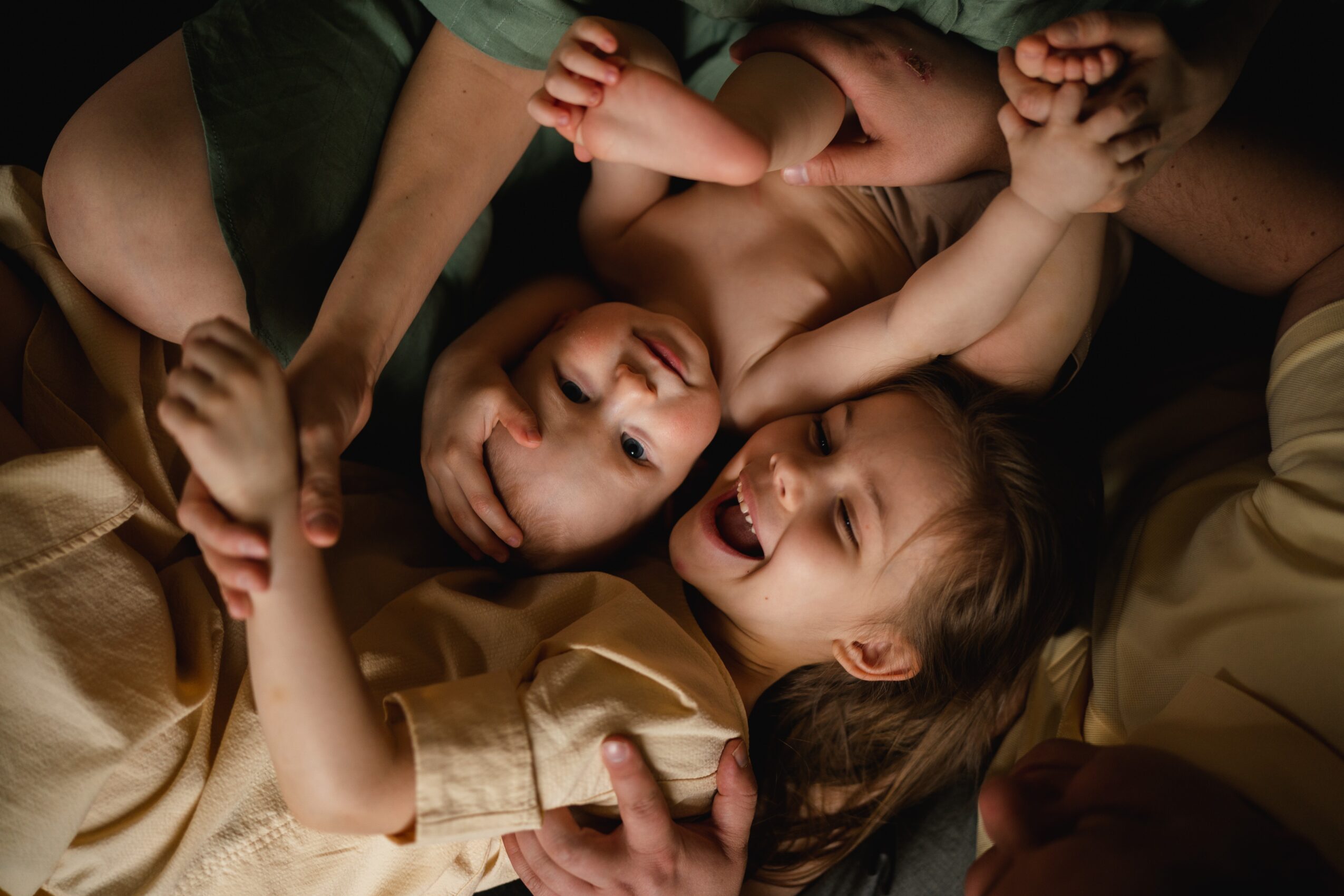 Children lying on the floor, smiling and playing together, with arms intertwined and heads close. Bright, joyful scene.