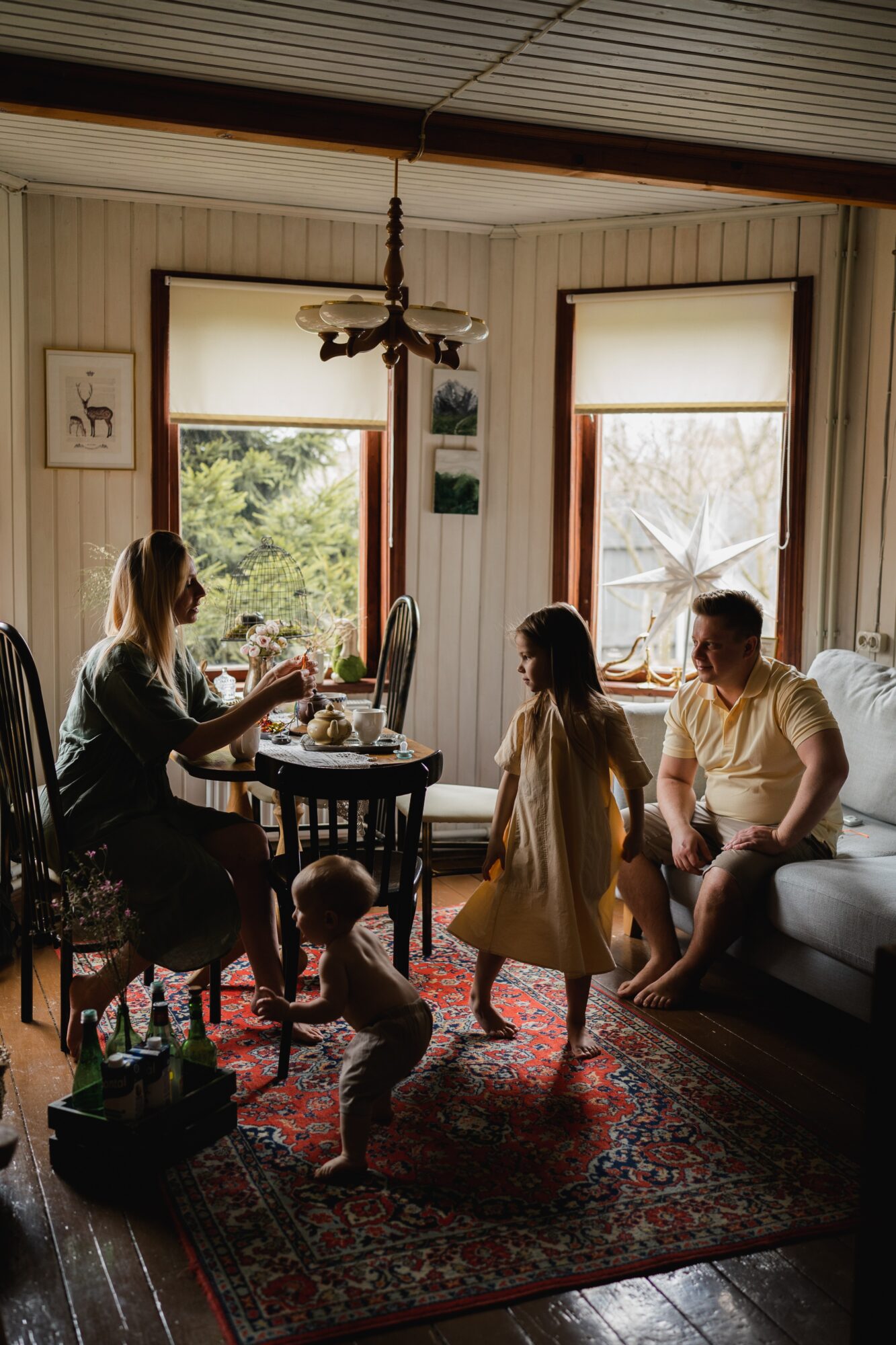 Family of four in a living room with large windows, two children standing, two adults sitting, rug on wooden floor.