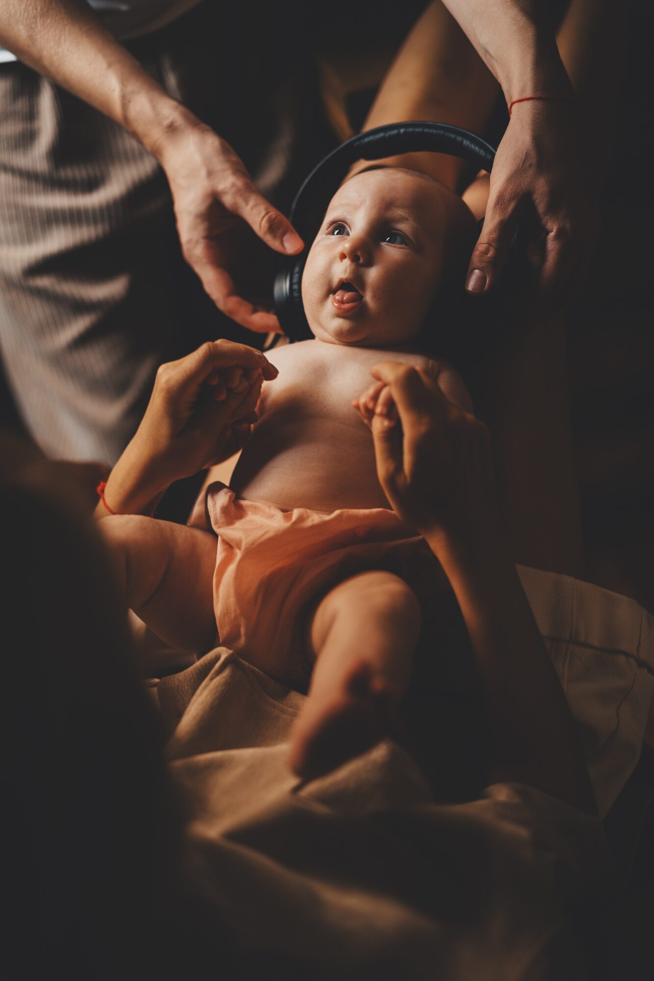 Baby lying on a surface, looking up with adults' hands adjusting a headband, warm lighting, focused on baby's face.