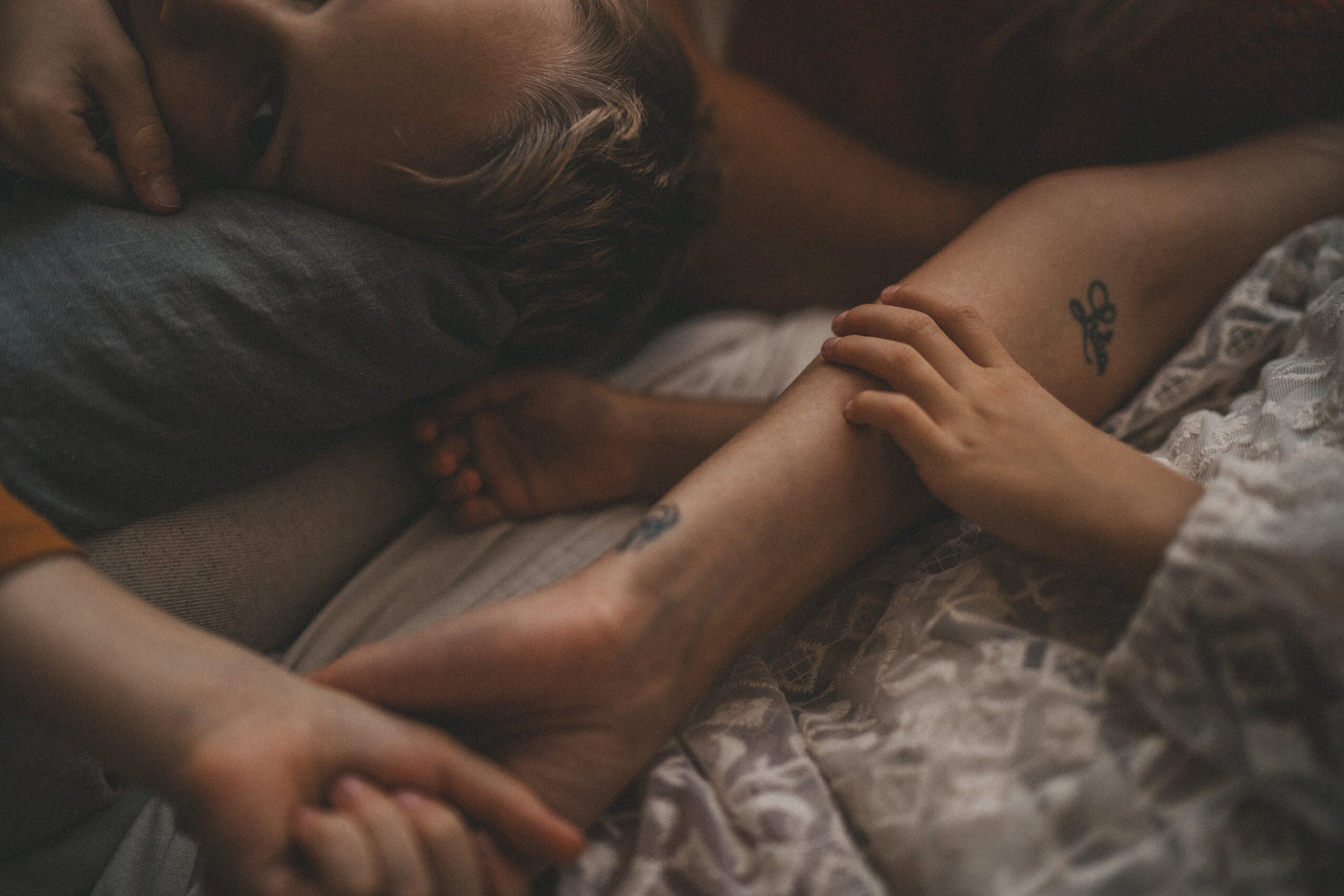 Person lying down with arm extended, holding another person's arm, in a dimly lit setting.