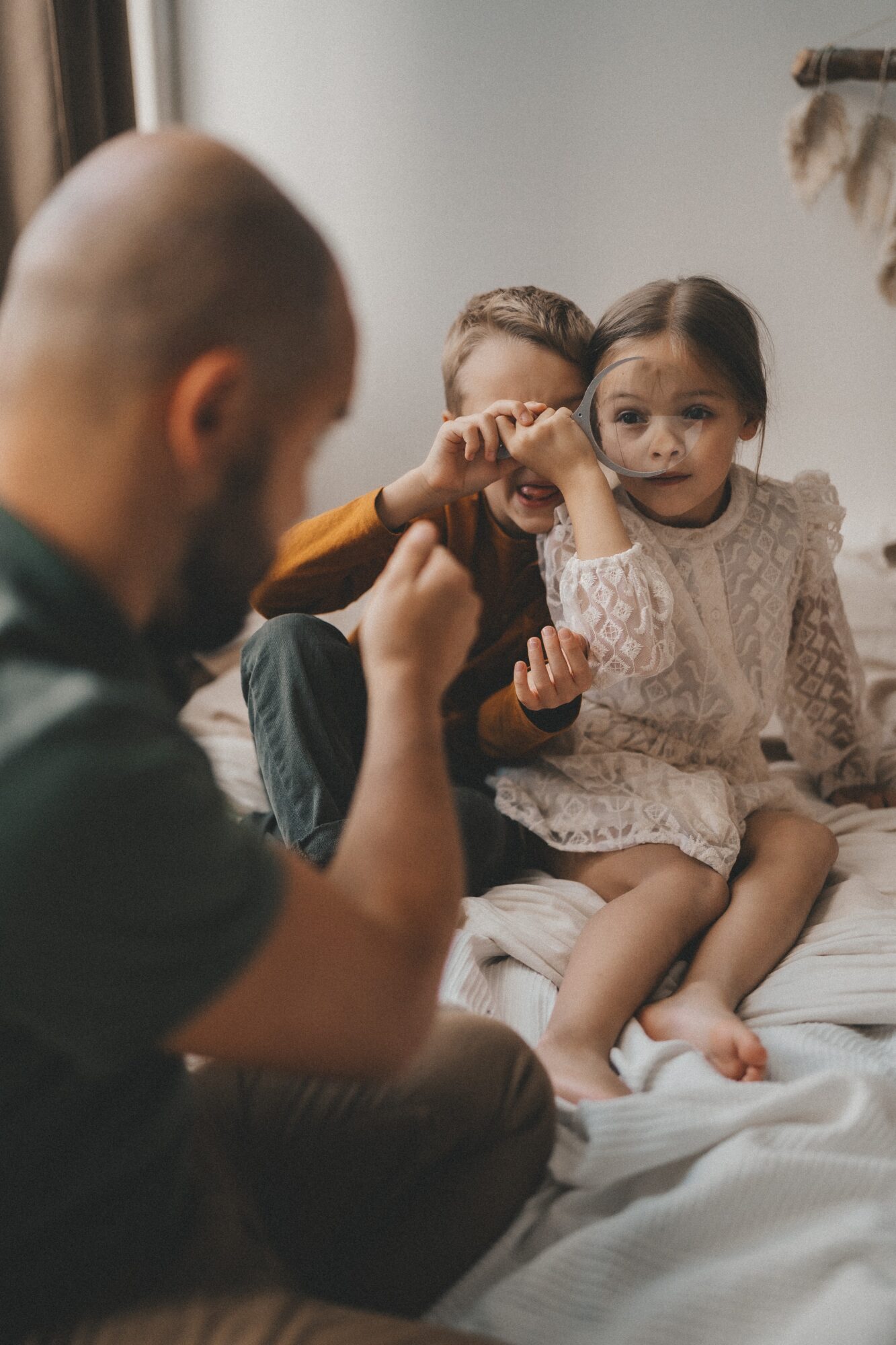 A man and two young girls sitting on a bed indoors, with the man showing something to the girls. One girl is holding her hand up to her face.