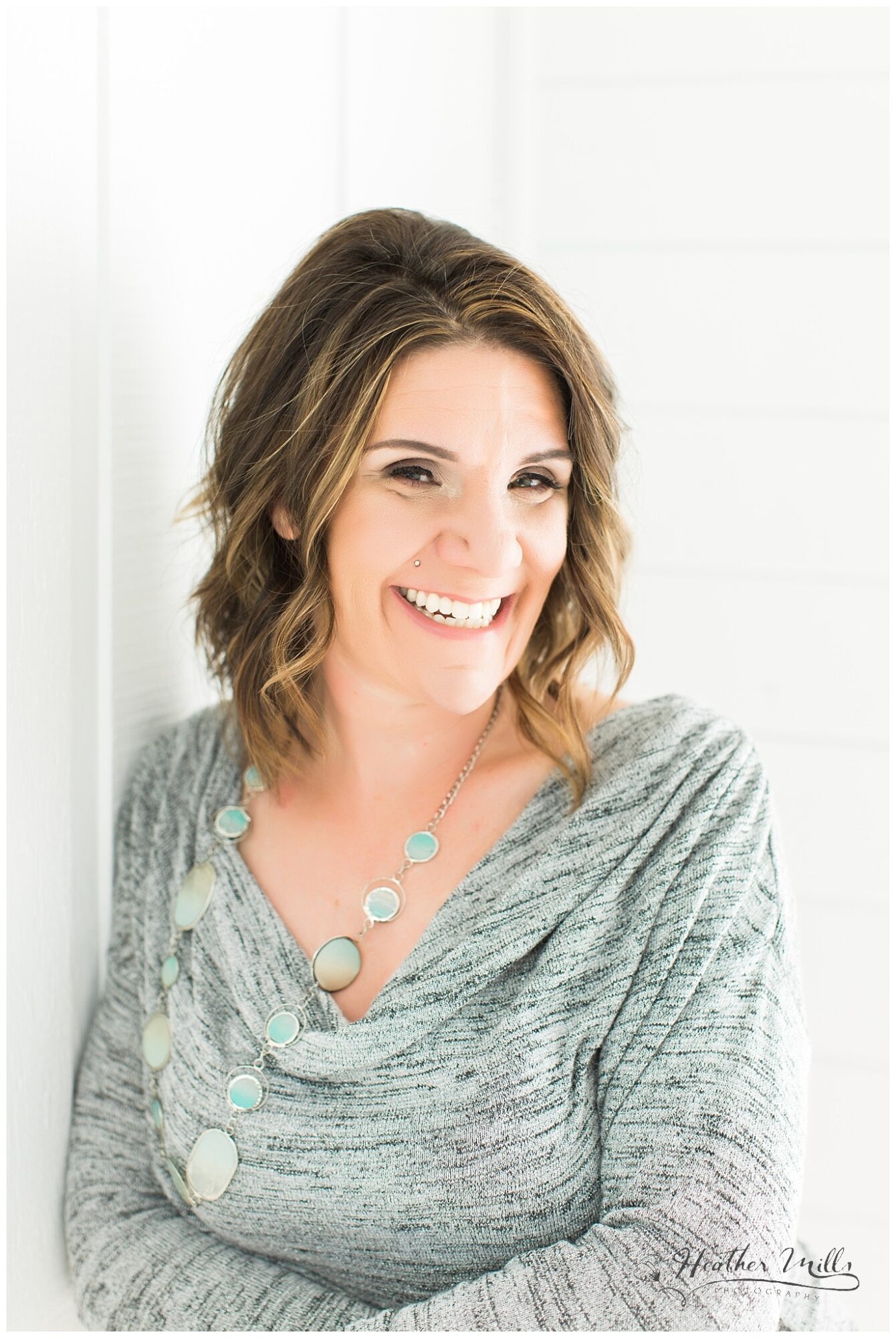 Woman with shoulder-length wavy hair smiling, wearing a gray top and a beaded necklace, standing against a white background.