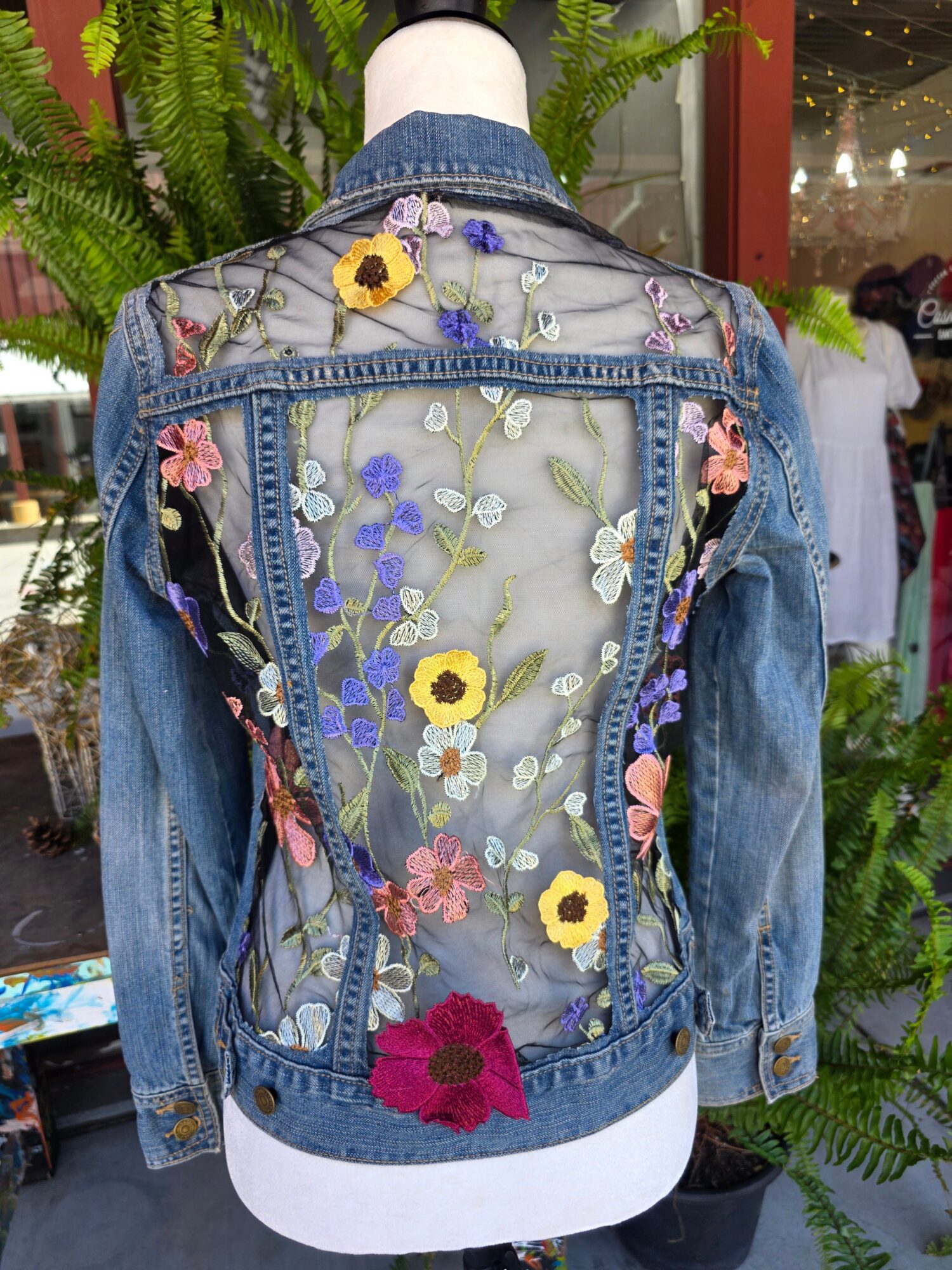 Back of a denim jacket with floral embroidery and sheer panel, displayed outdoors with plants and a store window behind.