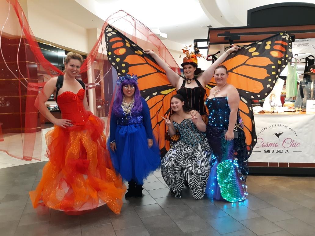 Five women in colorful costumes, one with butterfly wings, posing indoors in a shopping mall.