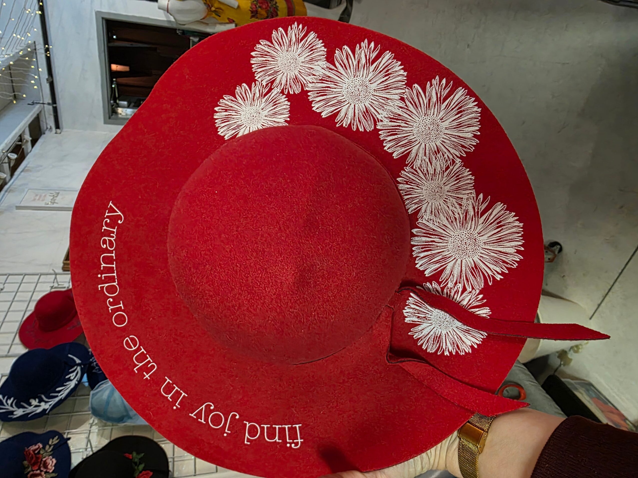 Red wide-brimmed hat with white floral design and embroidered text, held by a person. Background shows store interior.