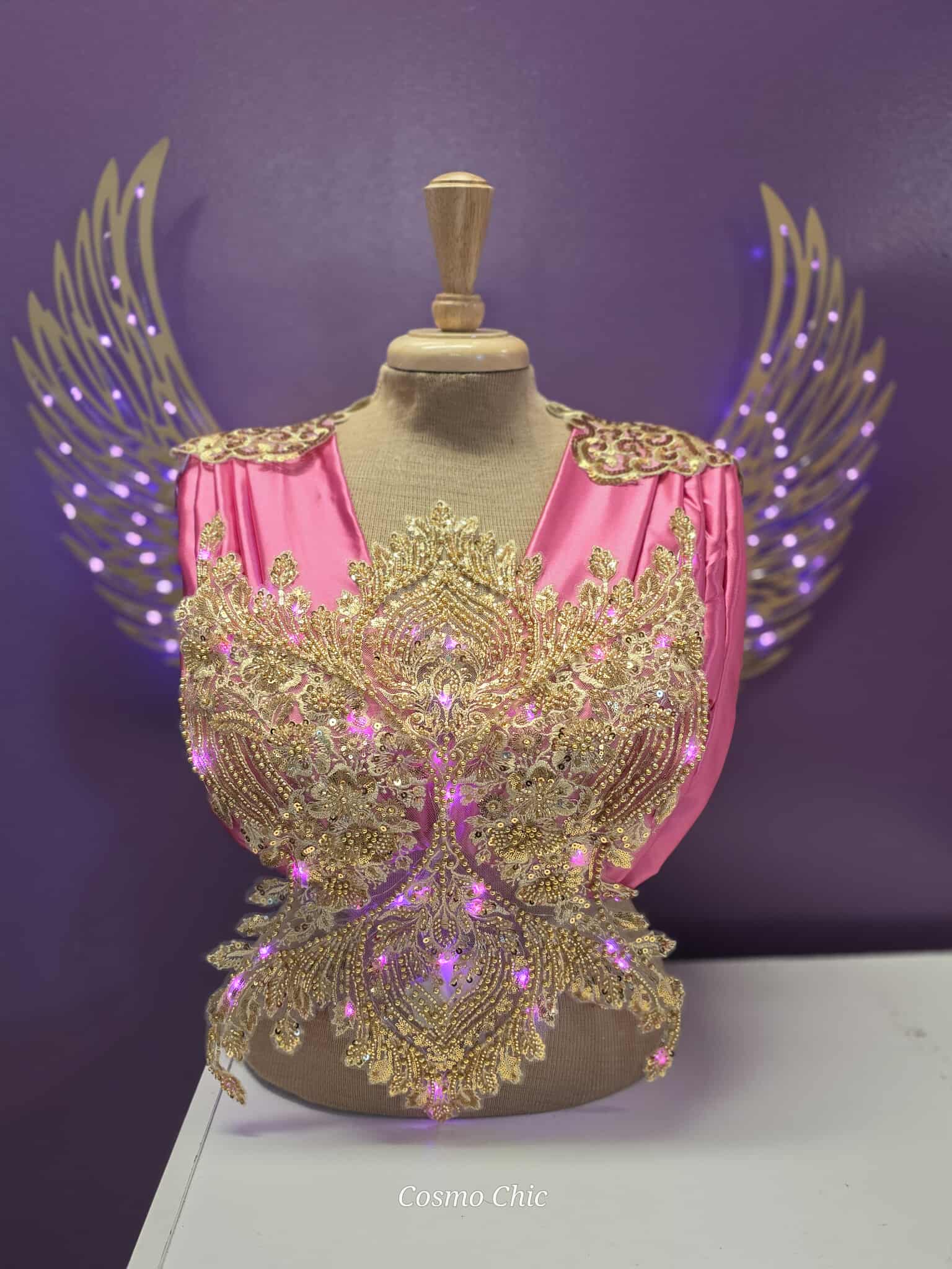 Display mannequin with pink and gold costume, gold wings, and purple background, labeled 'Cosmo Chic'.