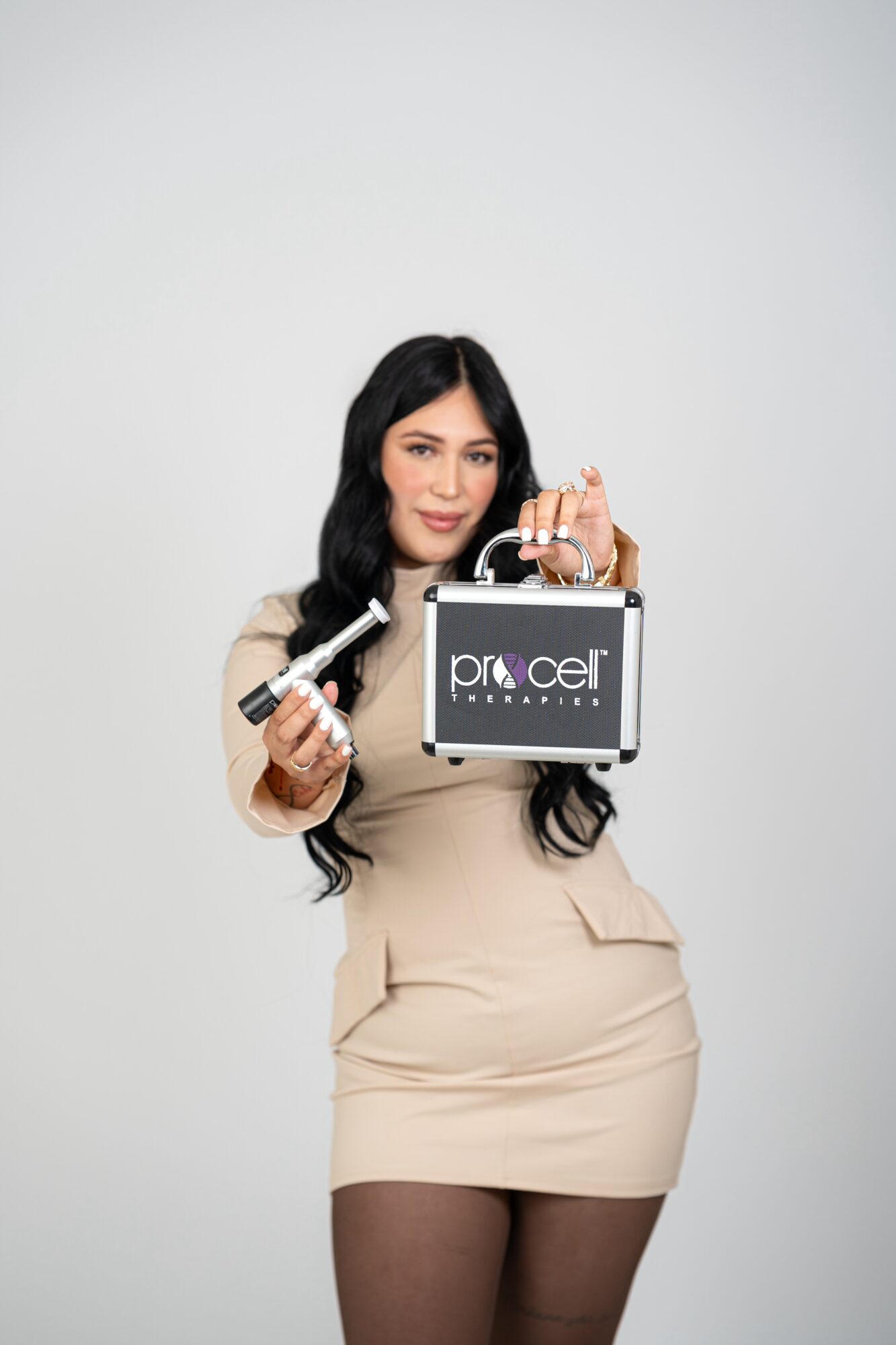 Woman with long dark hair holding beauty products and a black box, standing against a plain light background.