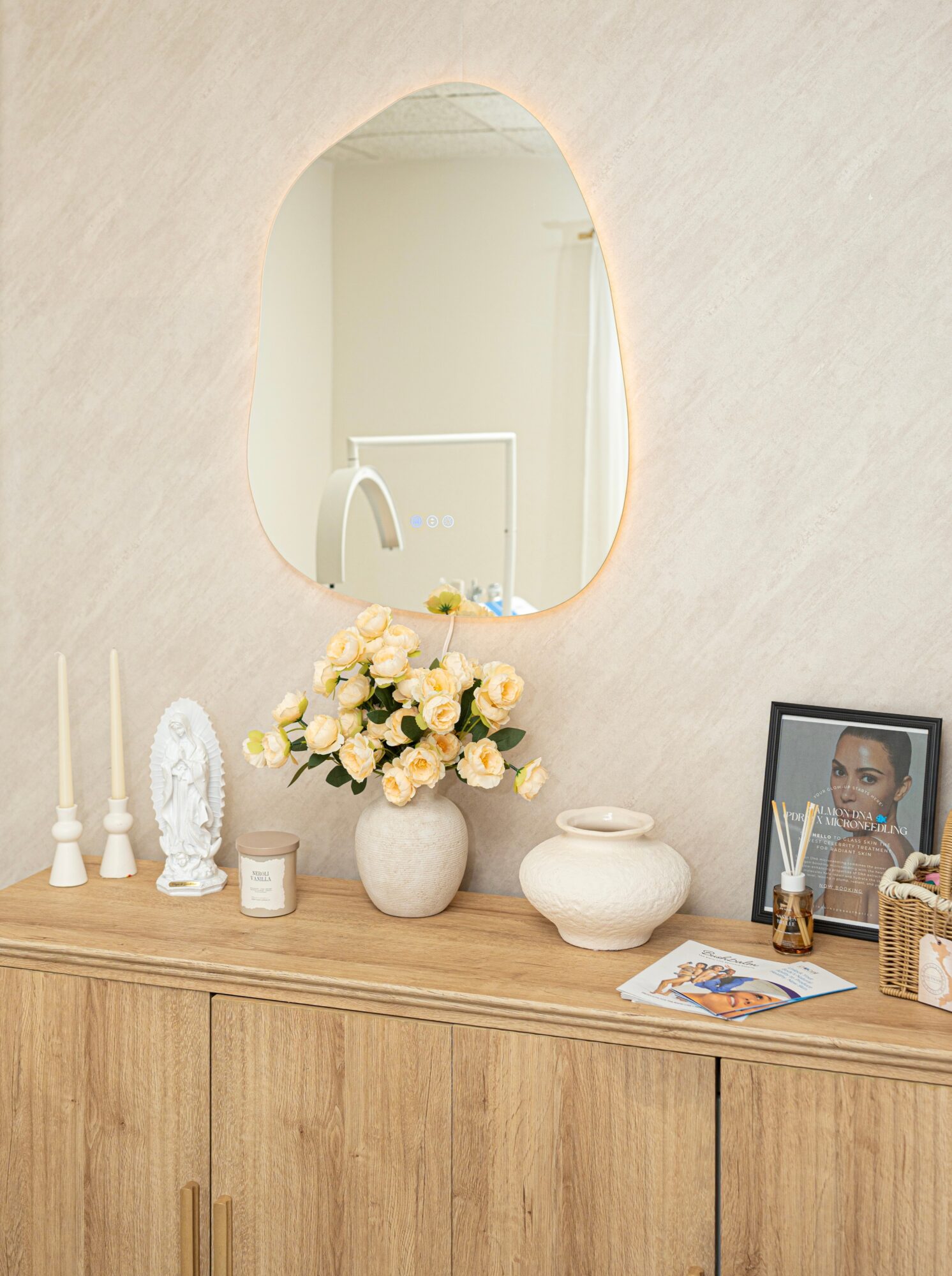 Decorative wooden sideboard with flowers, framed photo, candles, and decorative objects, reflected in an oval mirror on the wall.