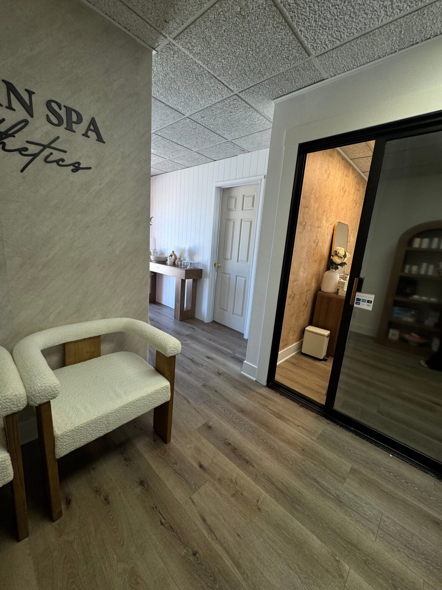 View of a hallway with a white chair, wooden floor, and a mirror reflecting a room with a beige wall and decorative items.