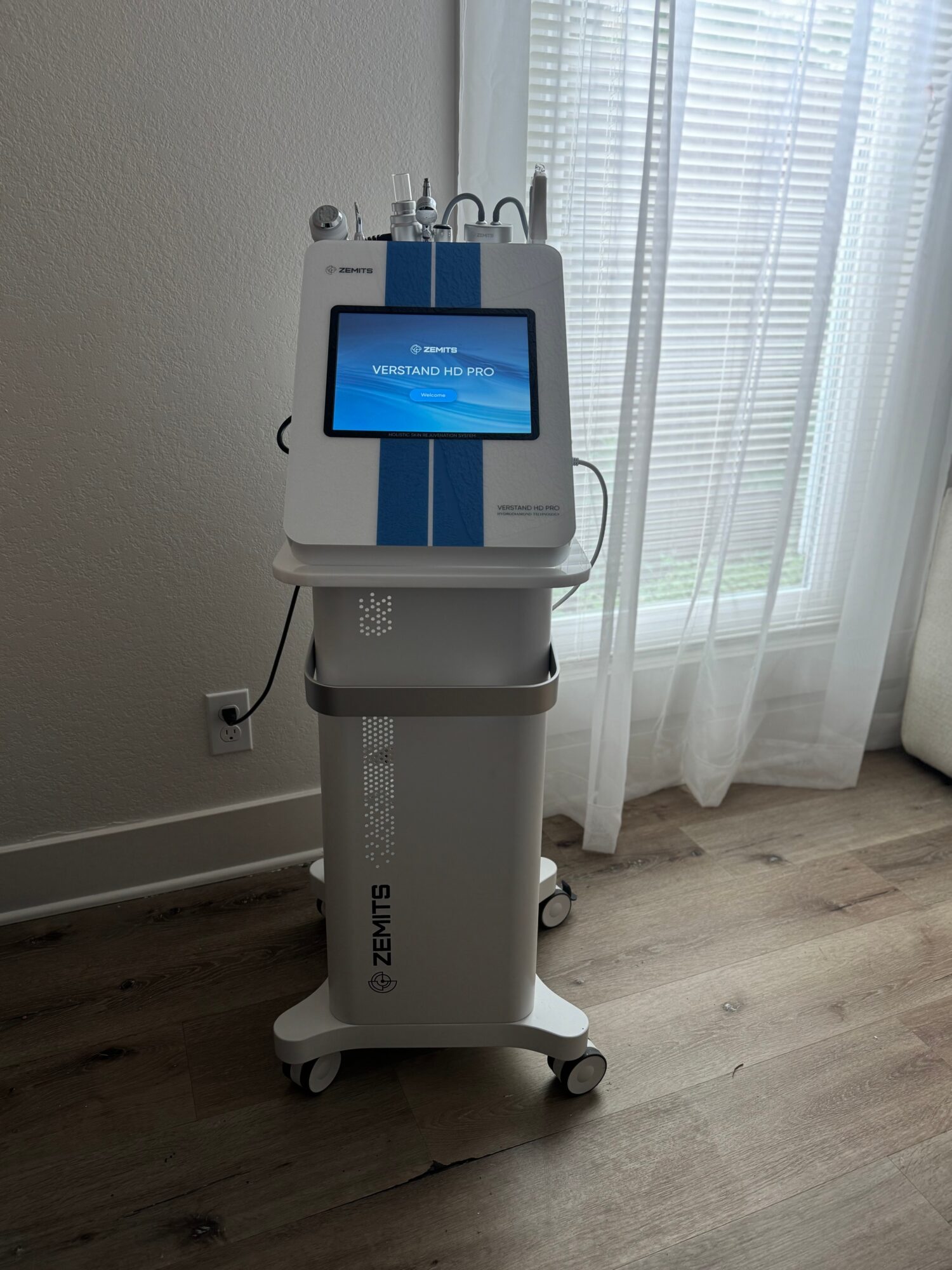 Medical device with a screen on a stand, positioned near a window with curtains, on a wooden floor.