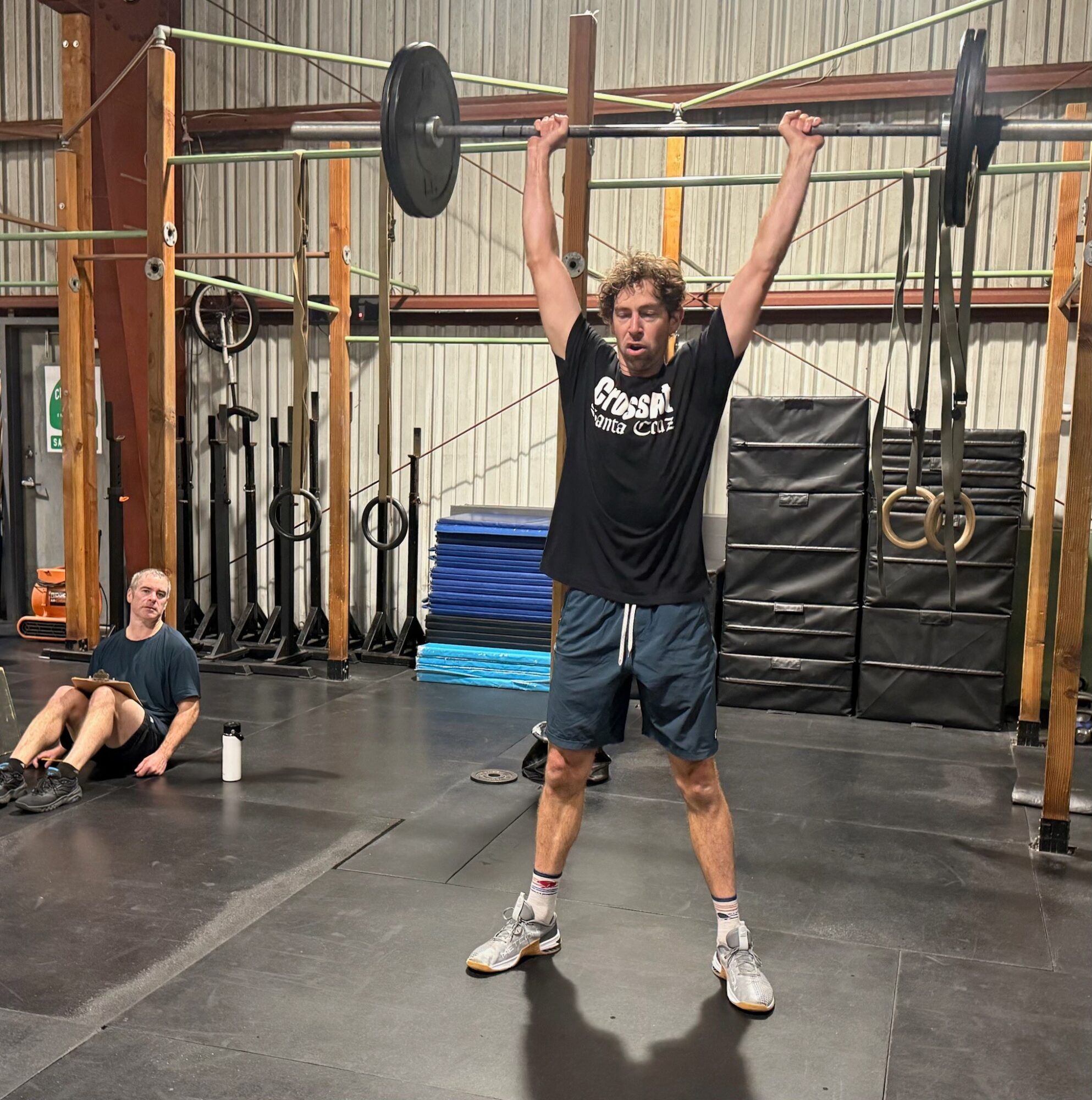 Young man lifting a barbell overhead in a gym with equipment and a seated person nearby.