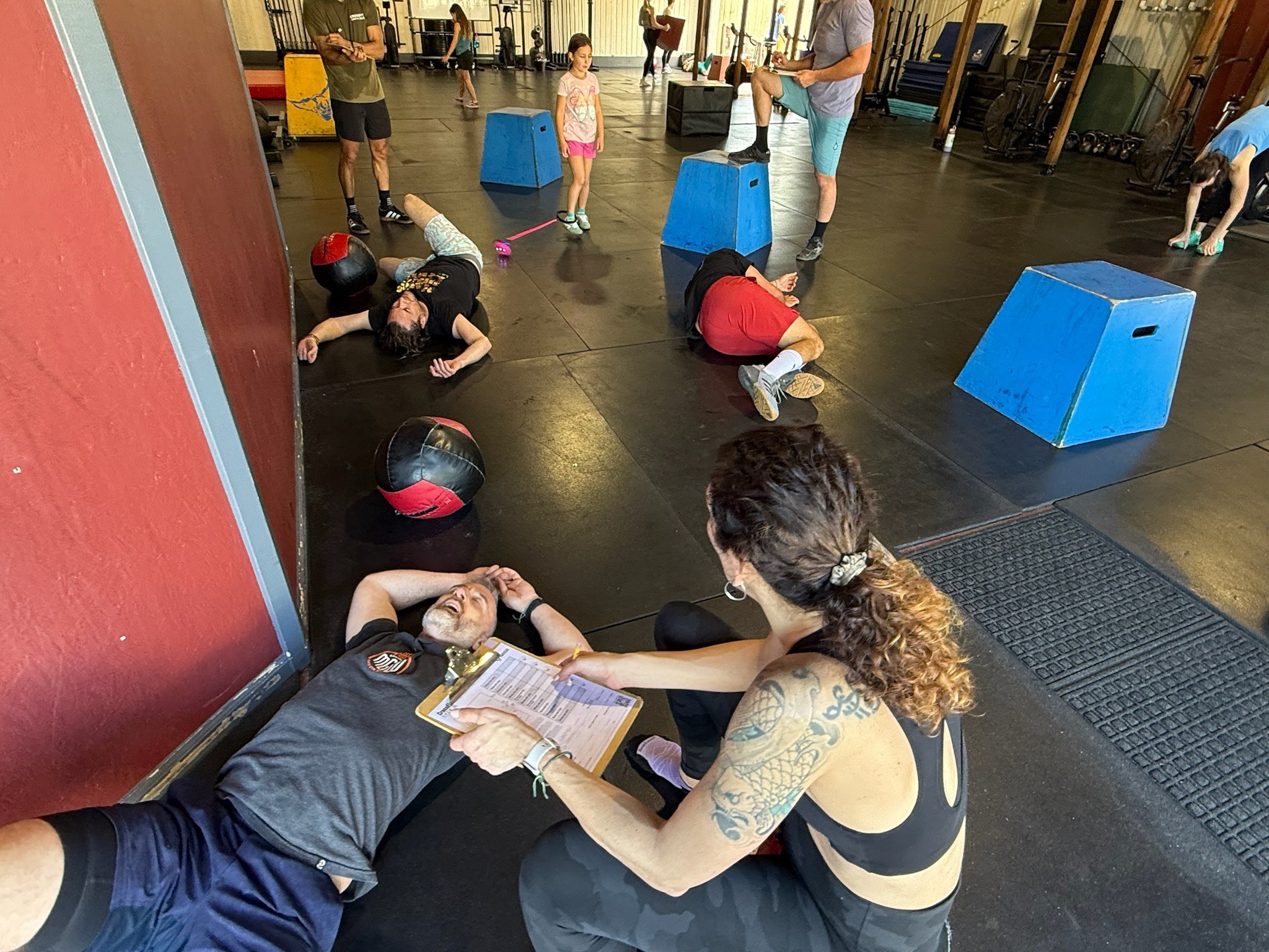 People participating in a fitness class, some lying on the floor, others using equipment, in a gym with large windows.
