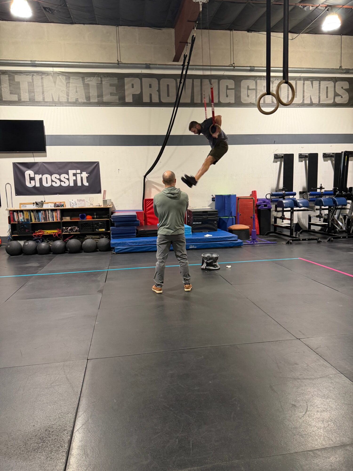 Person swinging on gym rings while another person watches in a CrossFit gym with equipment and mats.