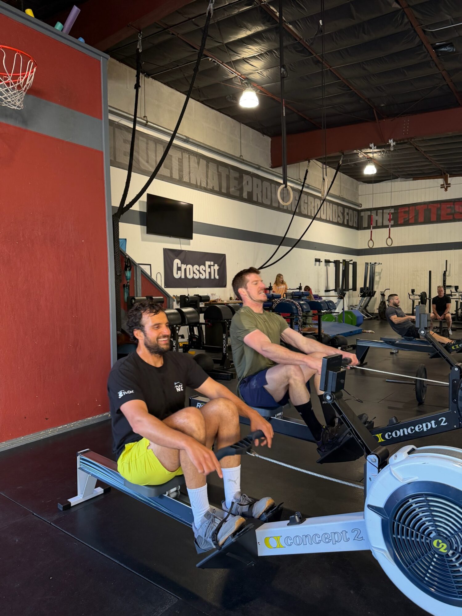 People using rowing machines in a gym with CrossFit signage, black ceiling, and bright lighting.