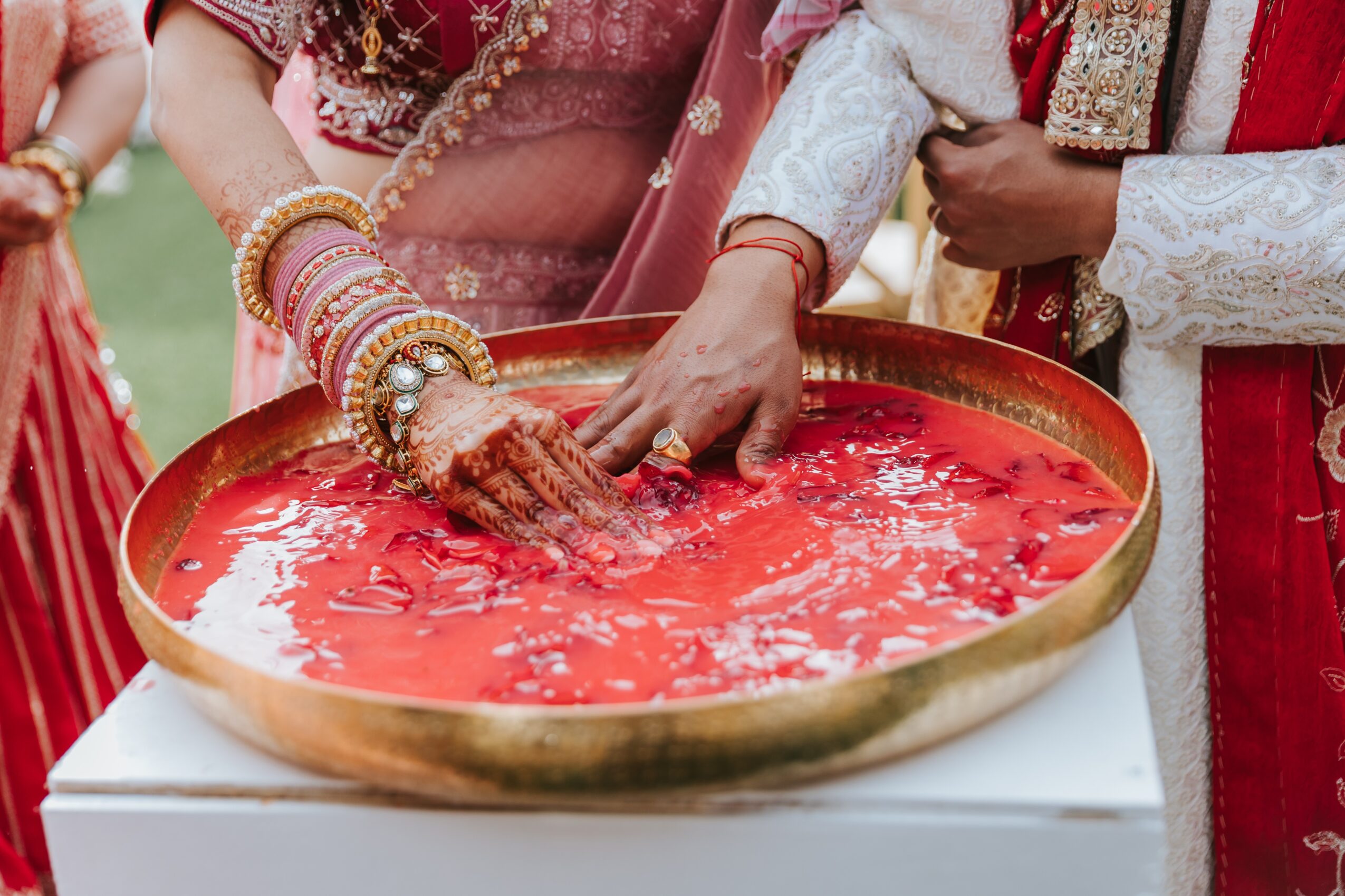 People participating in a traditional ceremony, with hands touching a large round tray filled with red liquid, wearing colorful clothing and jewelry.