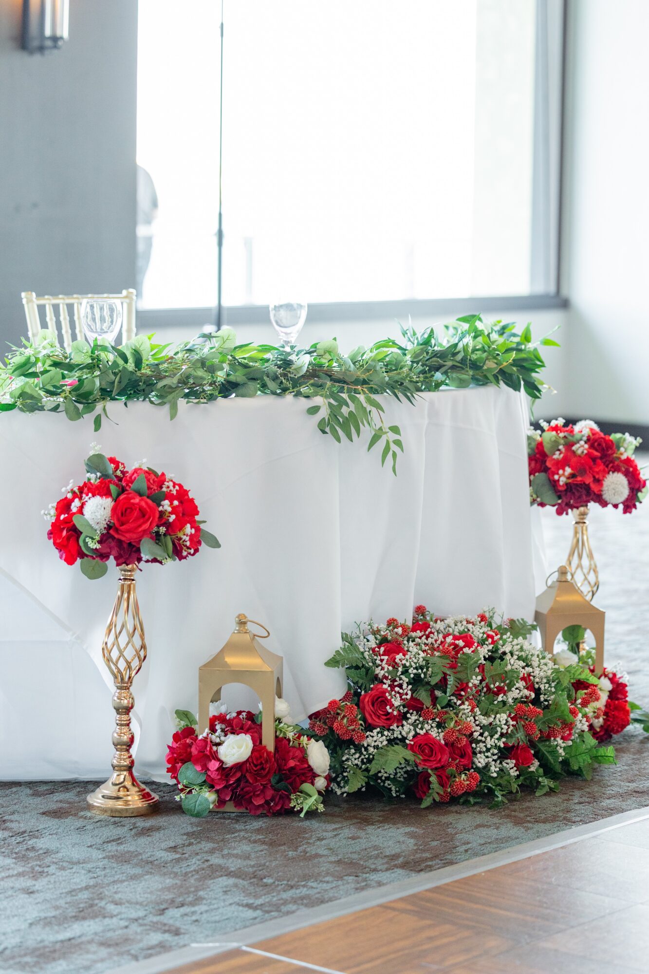 Floral arrangements with red, white, and pink flowers on gold stands and a white table with greenery, in a bright room.