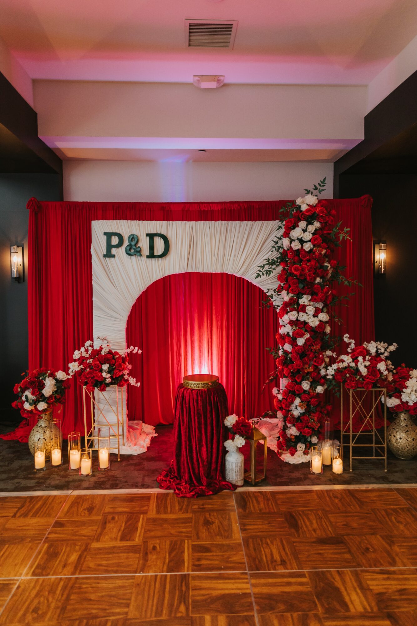 Decorative wedding backdrop with red curtains, floral arrangements, candles, and a small table, with initials P&D at the top.