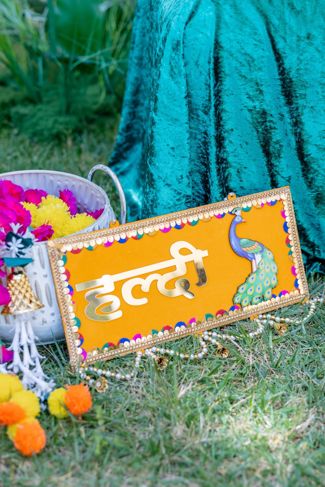Colorful sign with Hindi text and a peacock, decorated with flowers, placed on grass near a teal fabric backdrop.