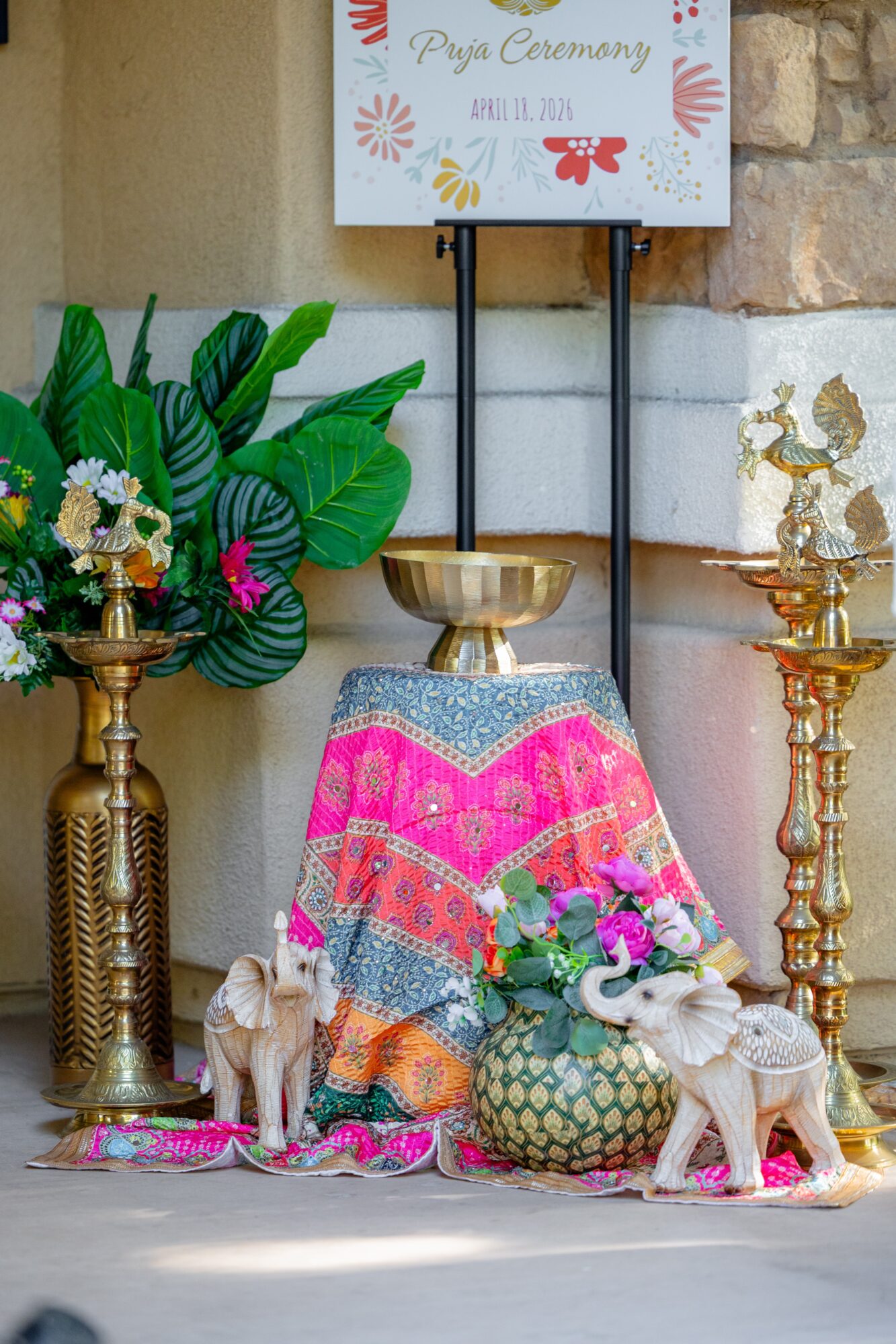 Decorative setup with plants, flowers, and animals, including two dogs, in front of a sign for a wedding ceremony, with a table covered in a colorful cloth.