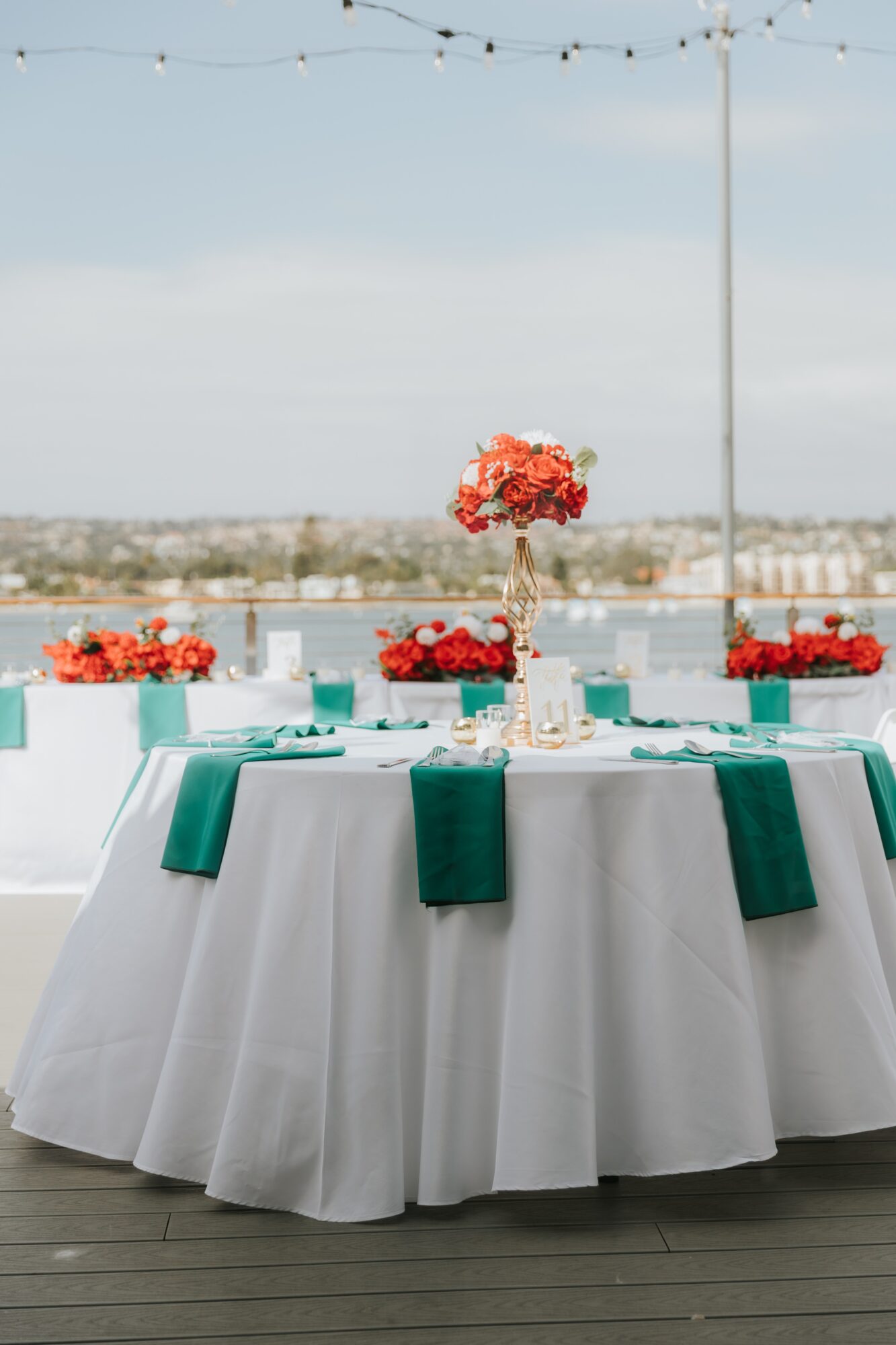 Round table with white tablecloth, teal napkins, and floral centerpieces outdoors under a string of lights, with a body of water in background.
