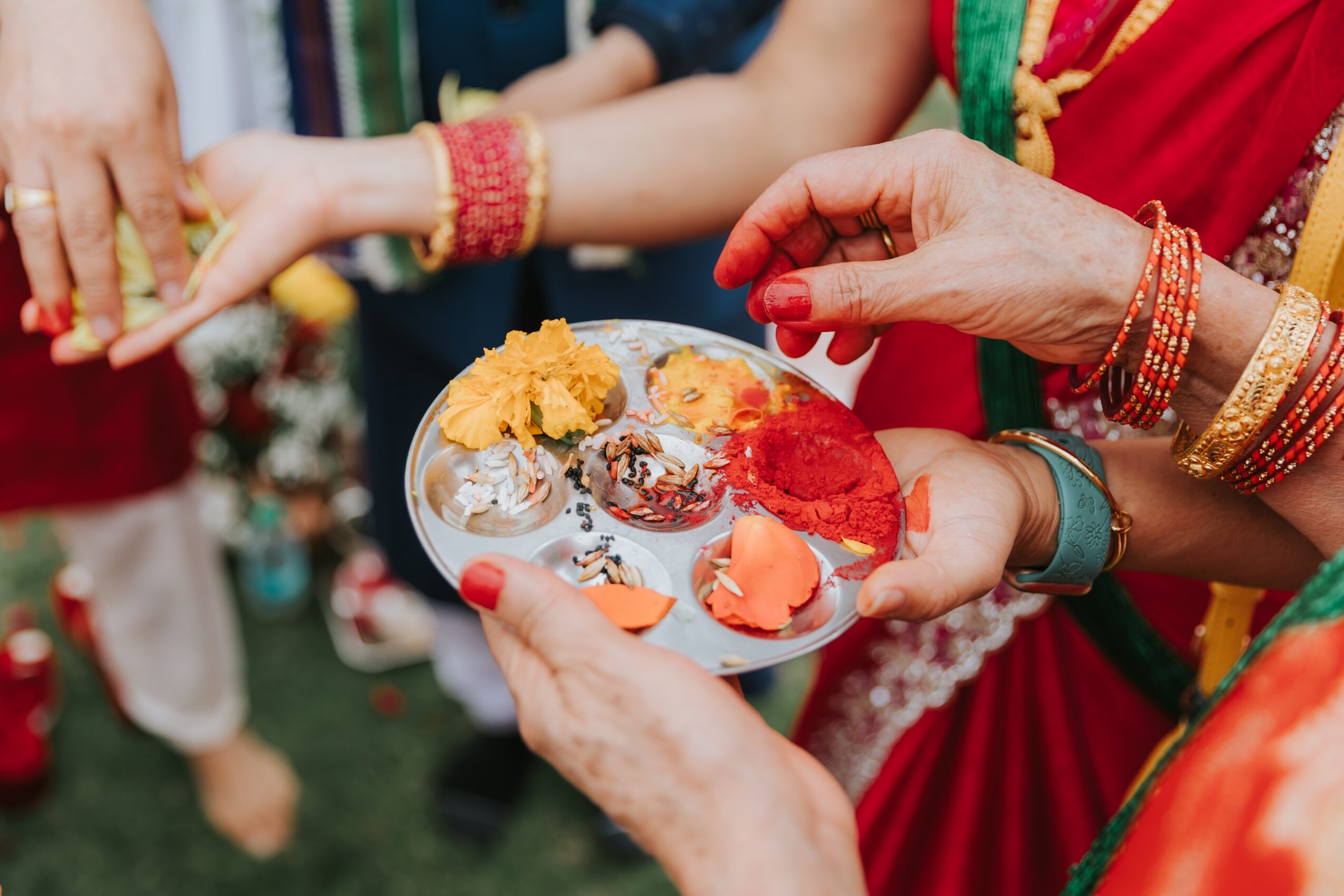 Person holding a plate with colorful sweets and flowers during a celebration, surrounded by others in traditional attire.