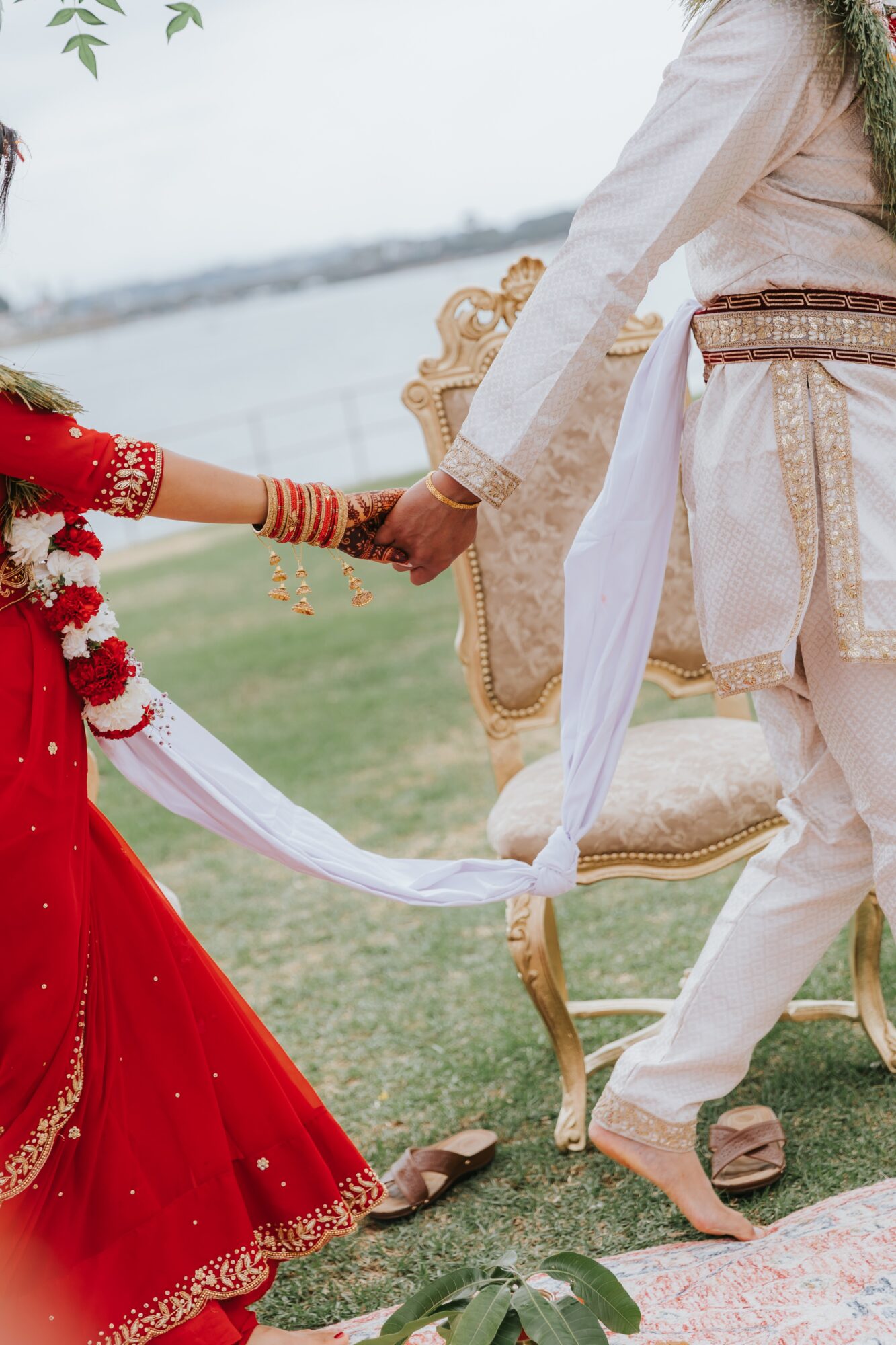 A couple holding hands outdoors, one in a red dress and the other in traditional attire, near a body of water.