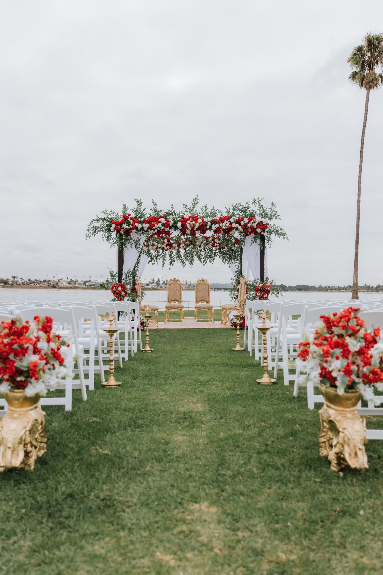Outdoor wedding setup with chairs, floral arch, and ocean view, under cloudy sky, with palm trees in background.