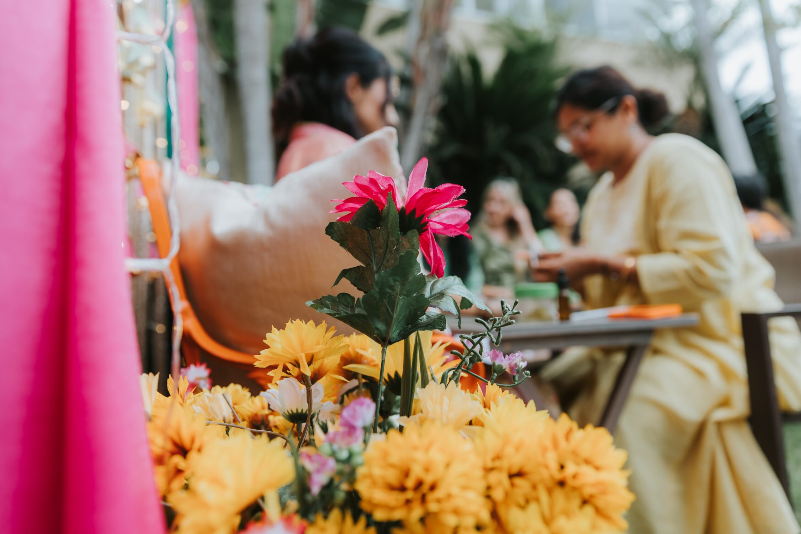 Flowers in foreground with people sitting and talking in background, outdoor setting, bright colors, casual atmosphere.