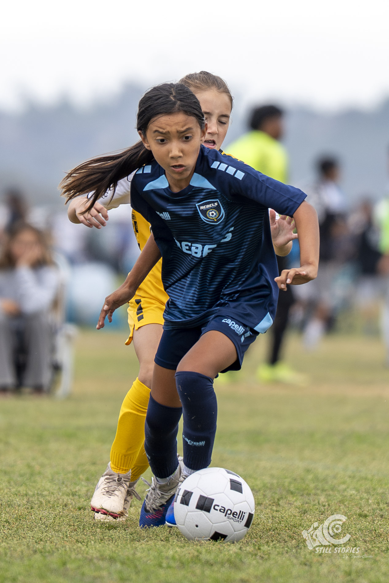 Two young girls playing soccer outdoors, one in a blue uniform chasing the ball, with spectators in the background.