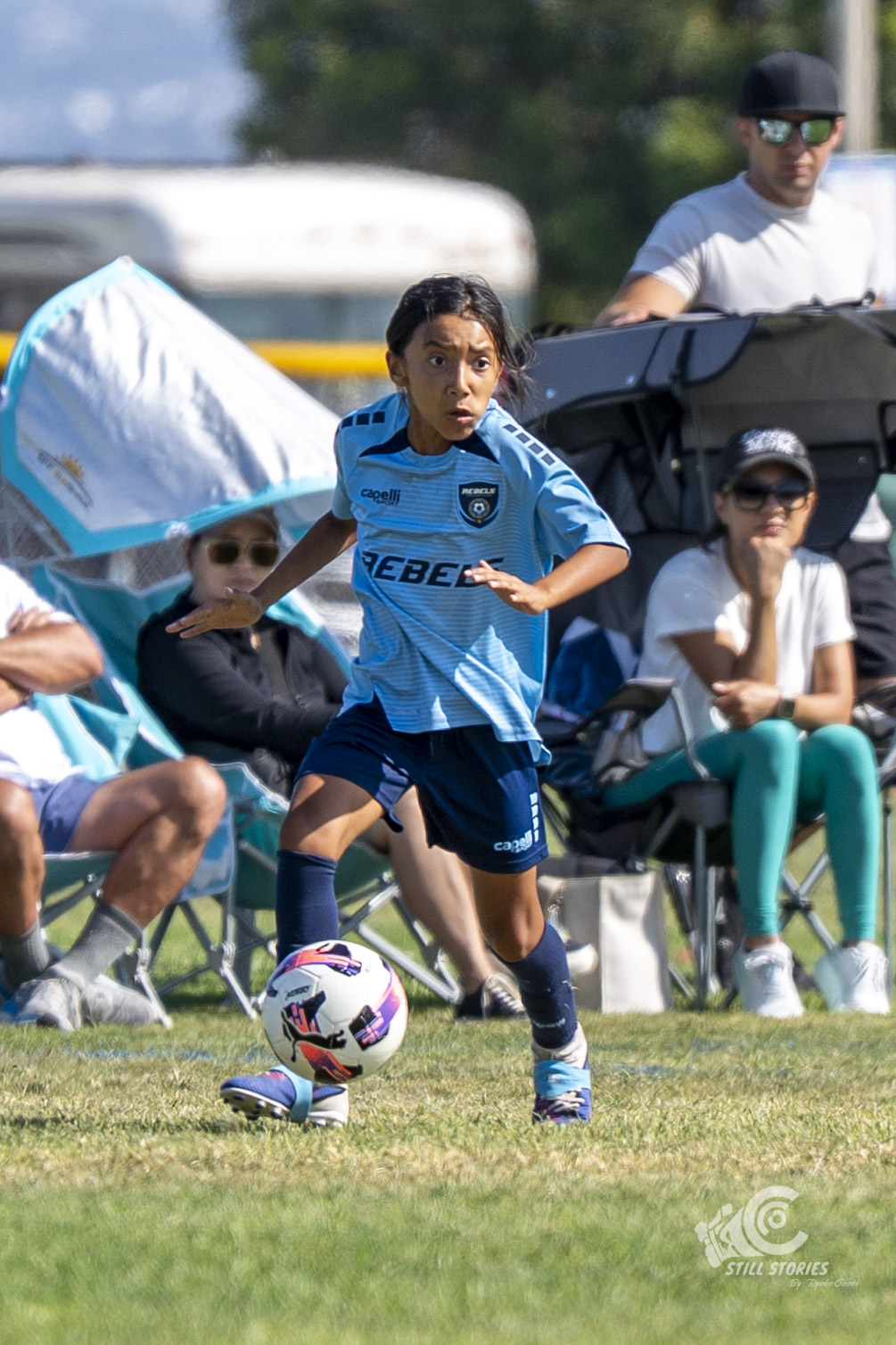 Young boy in blue soccer uniform dribbling a ball on a grassy field, with spectators sitting and standing in the background.
