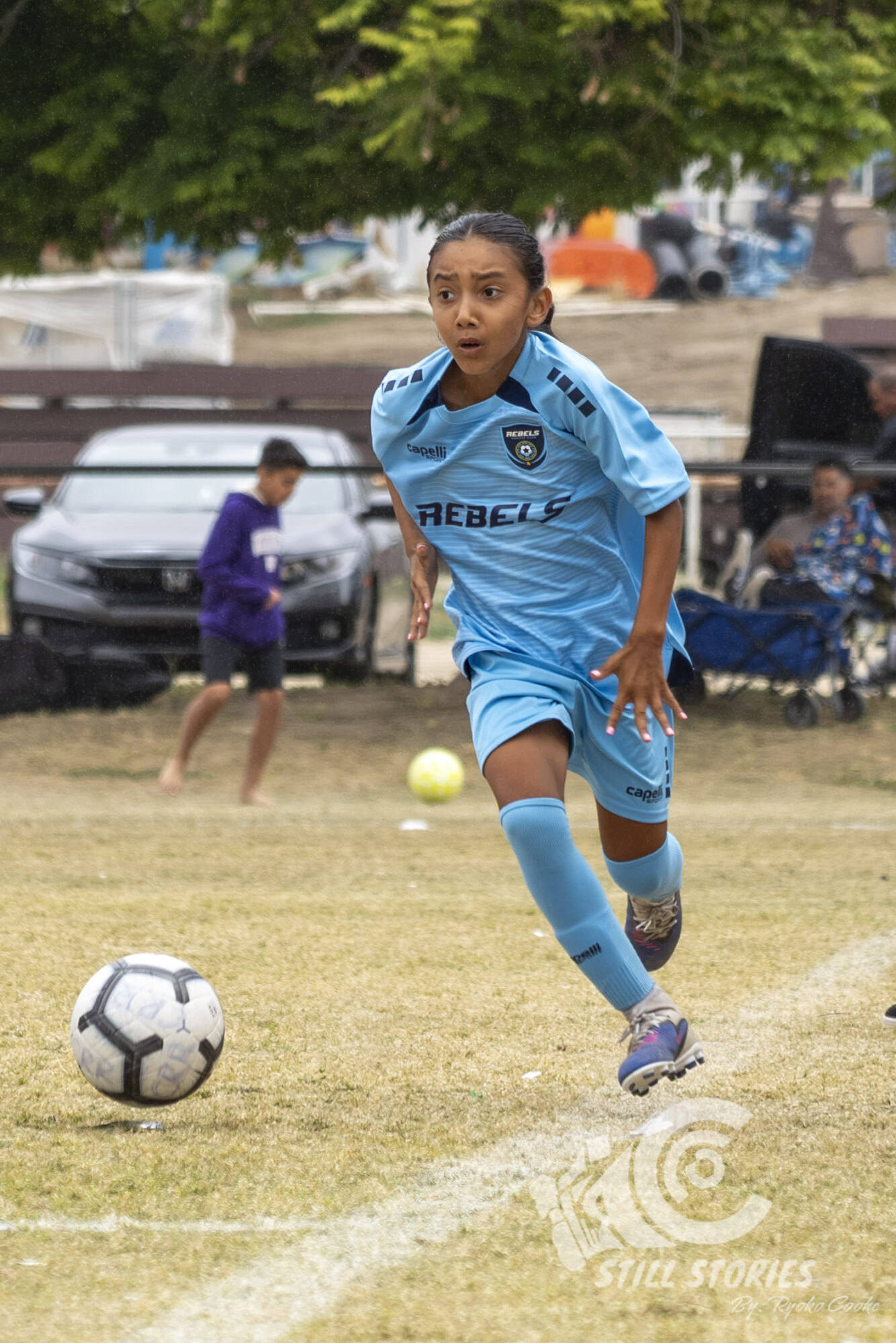 Young female soccer player in blue uniform running on field with soccer ball, trees, and people in background.