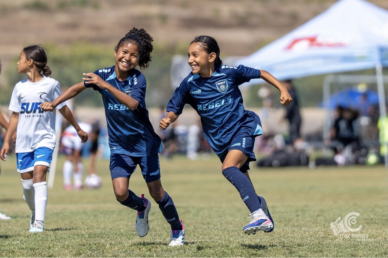 Three young girls in soccer uniforms running and smiling on a grassy field during a game.