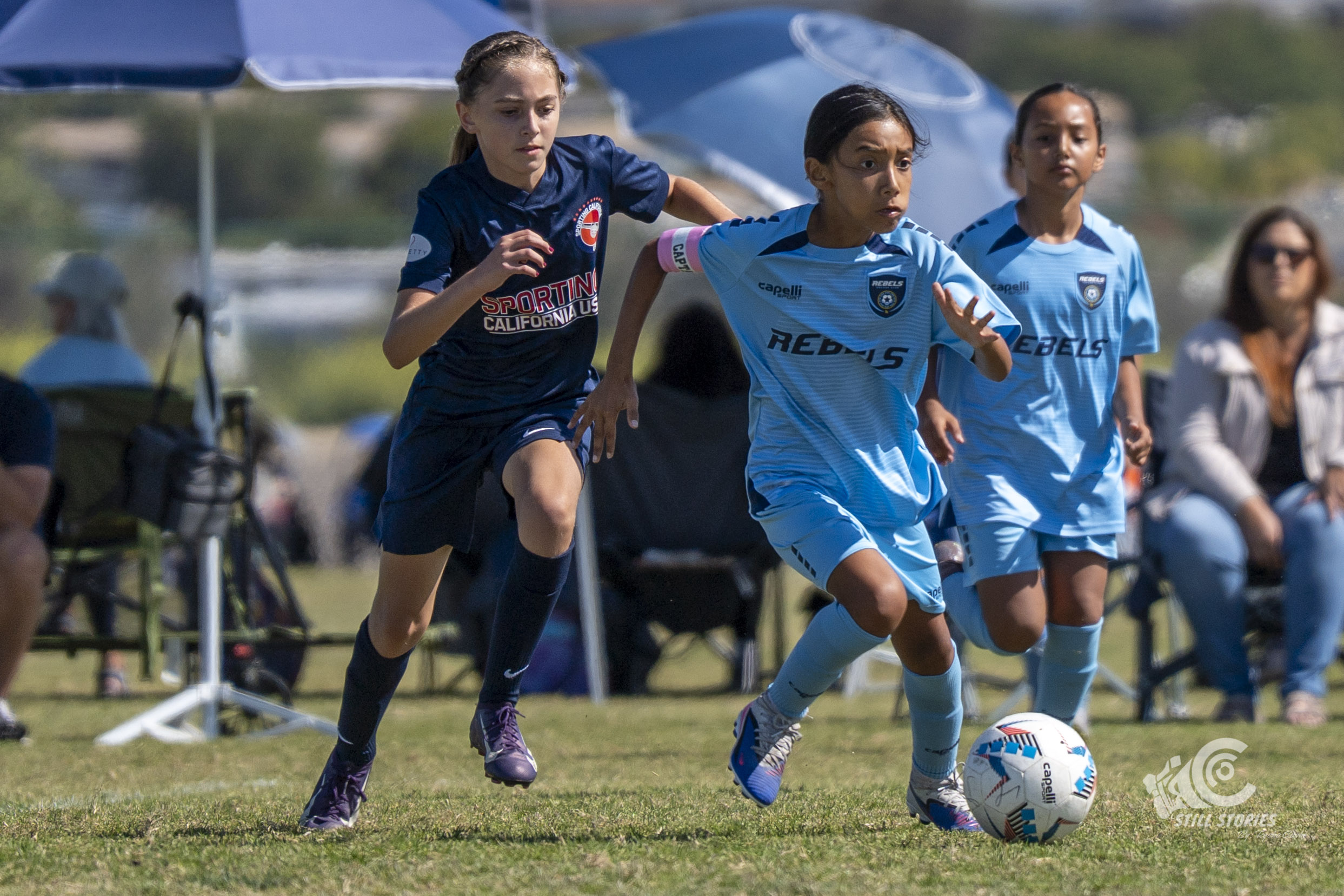 Three young female soccer players running on a field, with spectators sitting in the background under umbrellas.
