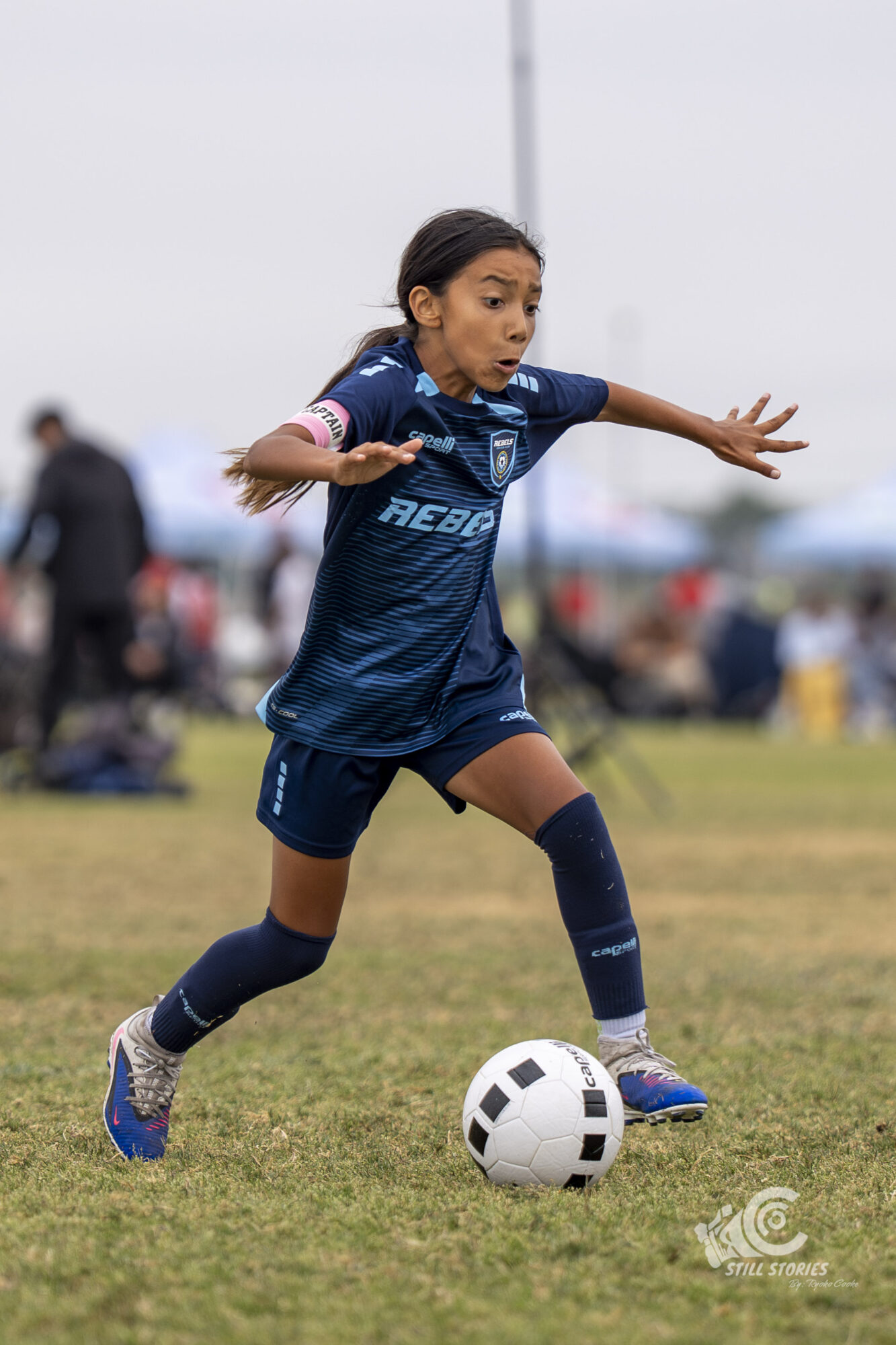 Young girl in a blue soccer uniform running on grass with a soccer ball, arms outstretched, focused on the game.