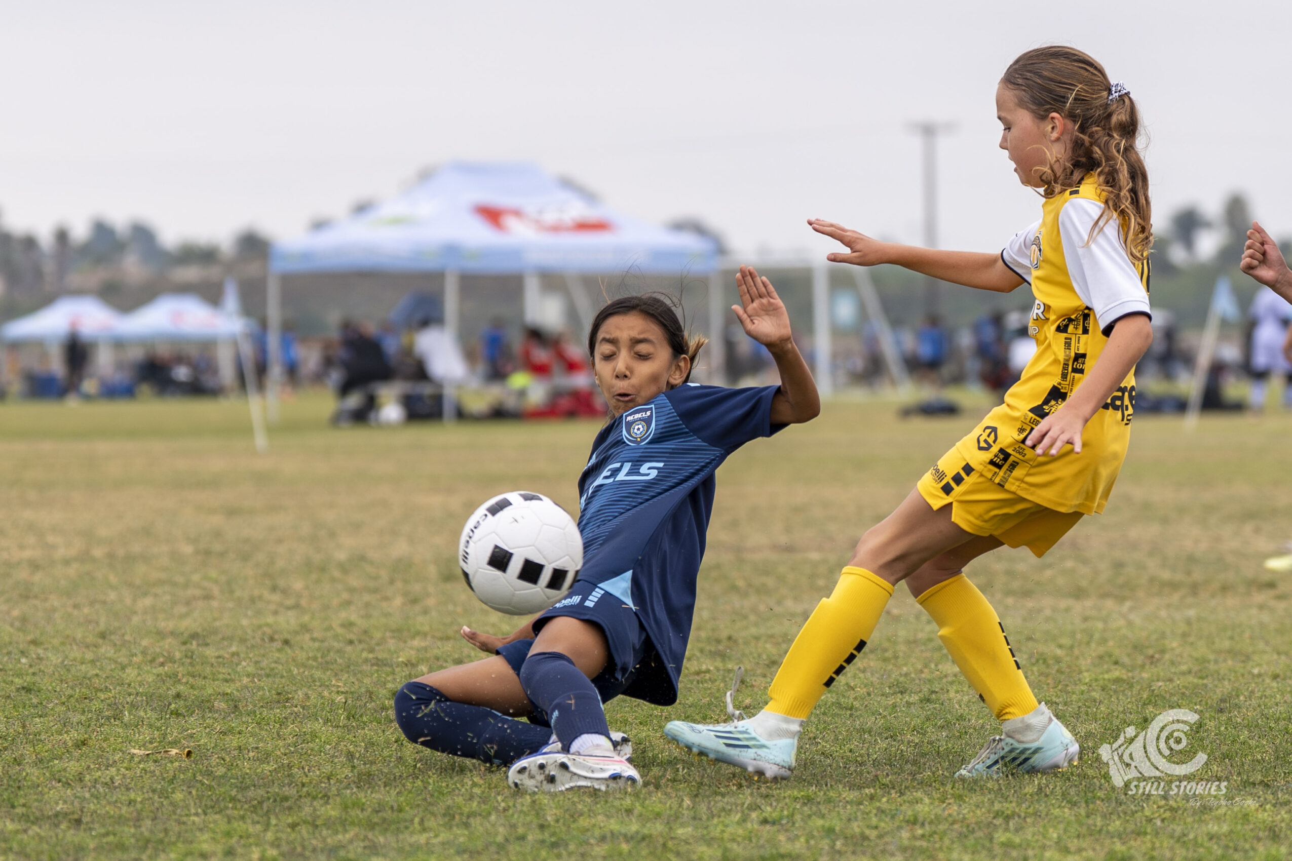 Two children playing soccer on a field, one in a blue jersey sliding to kick the ball, the other in yellow attempting to block.