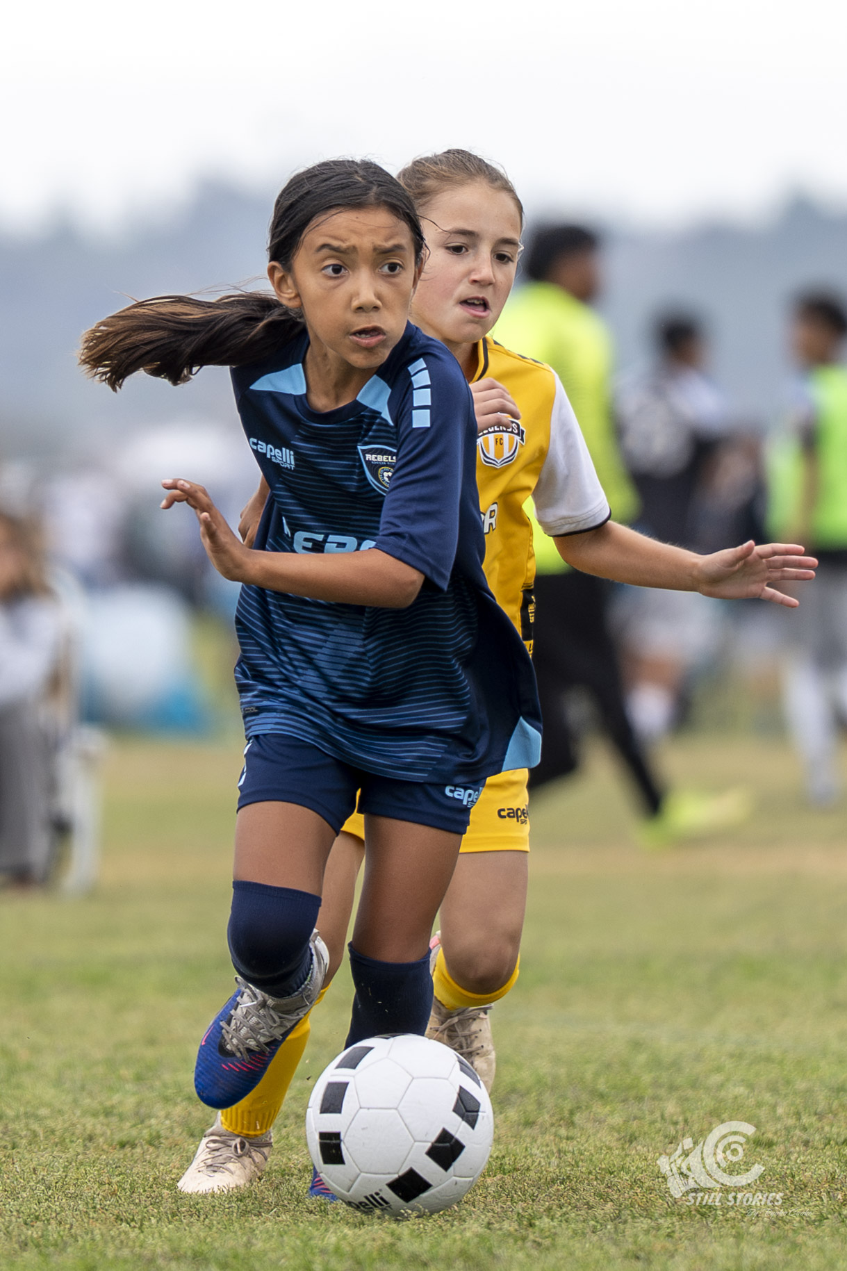 Two young girls playing soccer outdoors, one in a blue jersey and the other in a yellow jersey, chasing the ball.