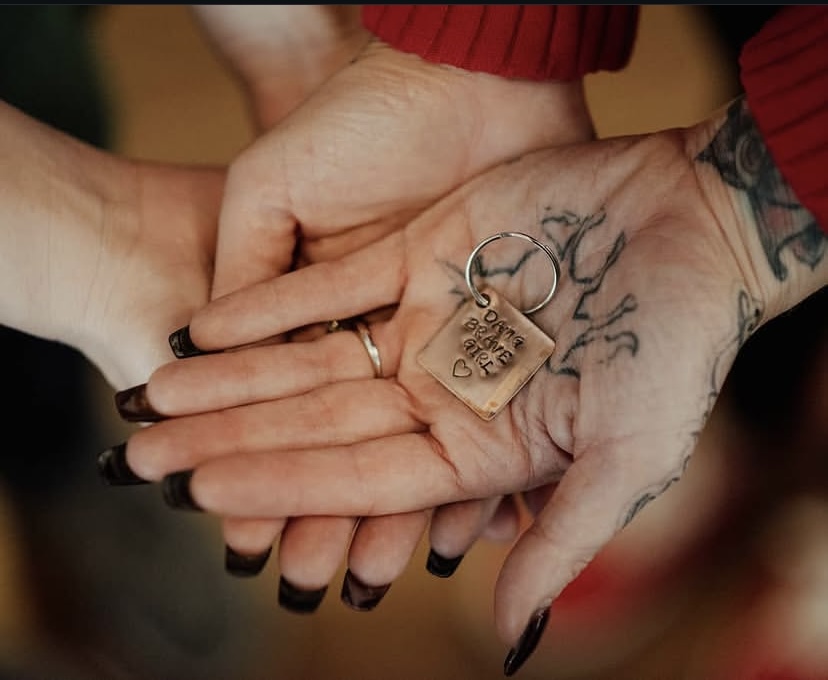 Two hands holding a keychain with a metal tag, one hand has tattoos and dark nail polish, the other hand is unadorned.